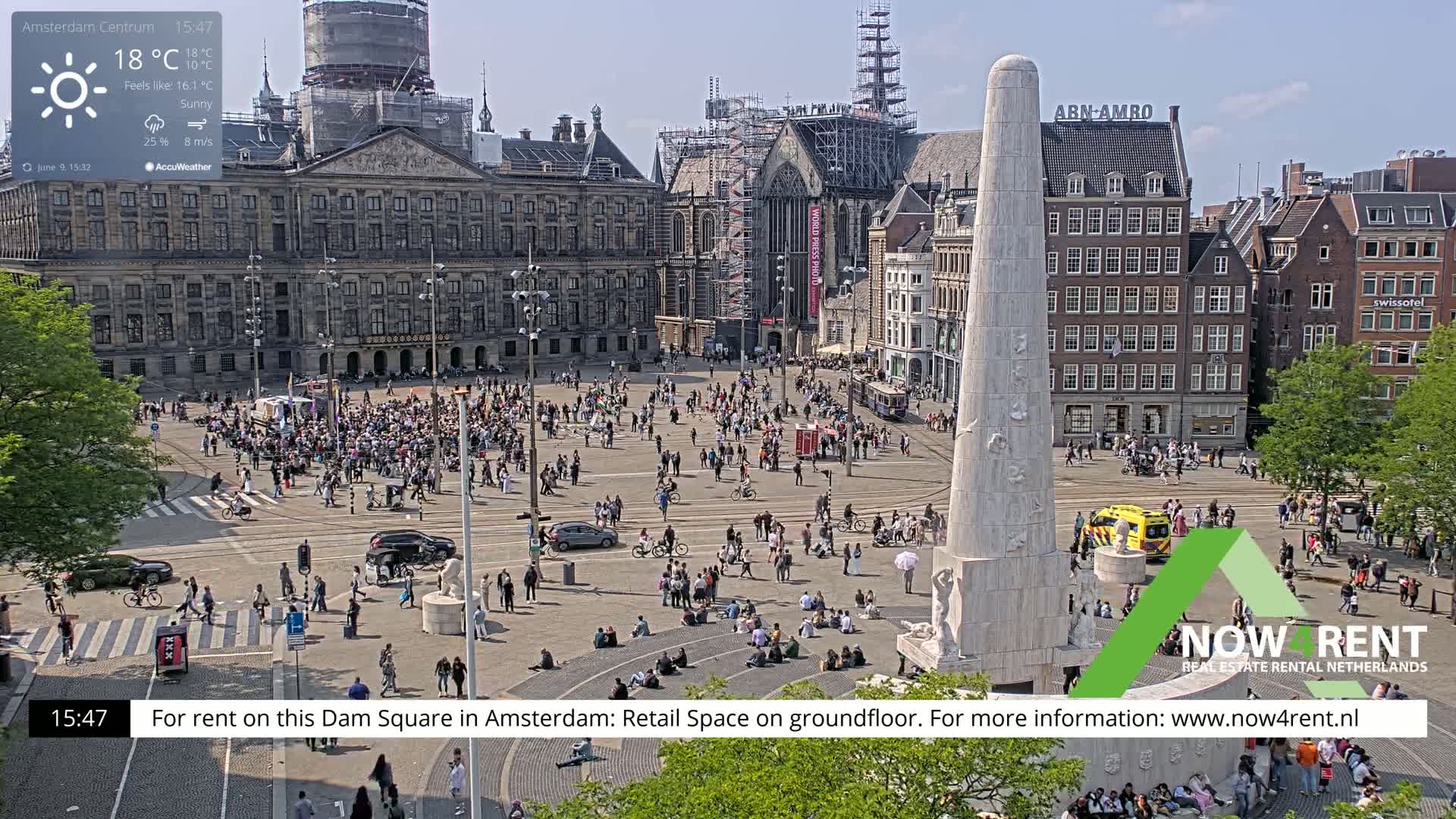 A sunny Dam Square in Amsterdam, populated with people, features a tall monument and several buildings, one of which is under construction.