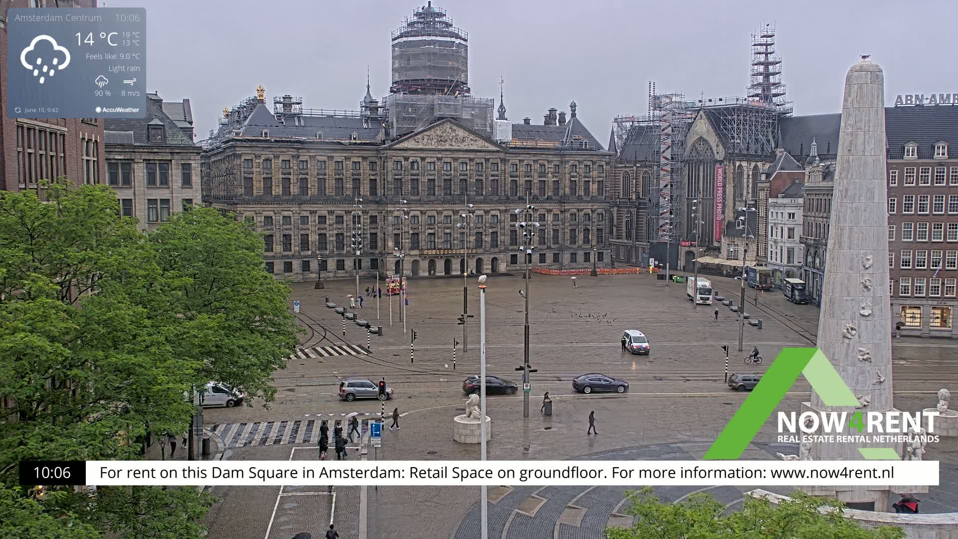A rainy view of Dam Square in Amsterdam, showing the Royal Palace under construction and a large obelisk.