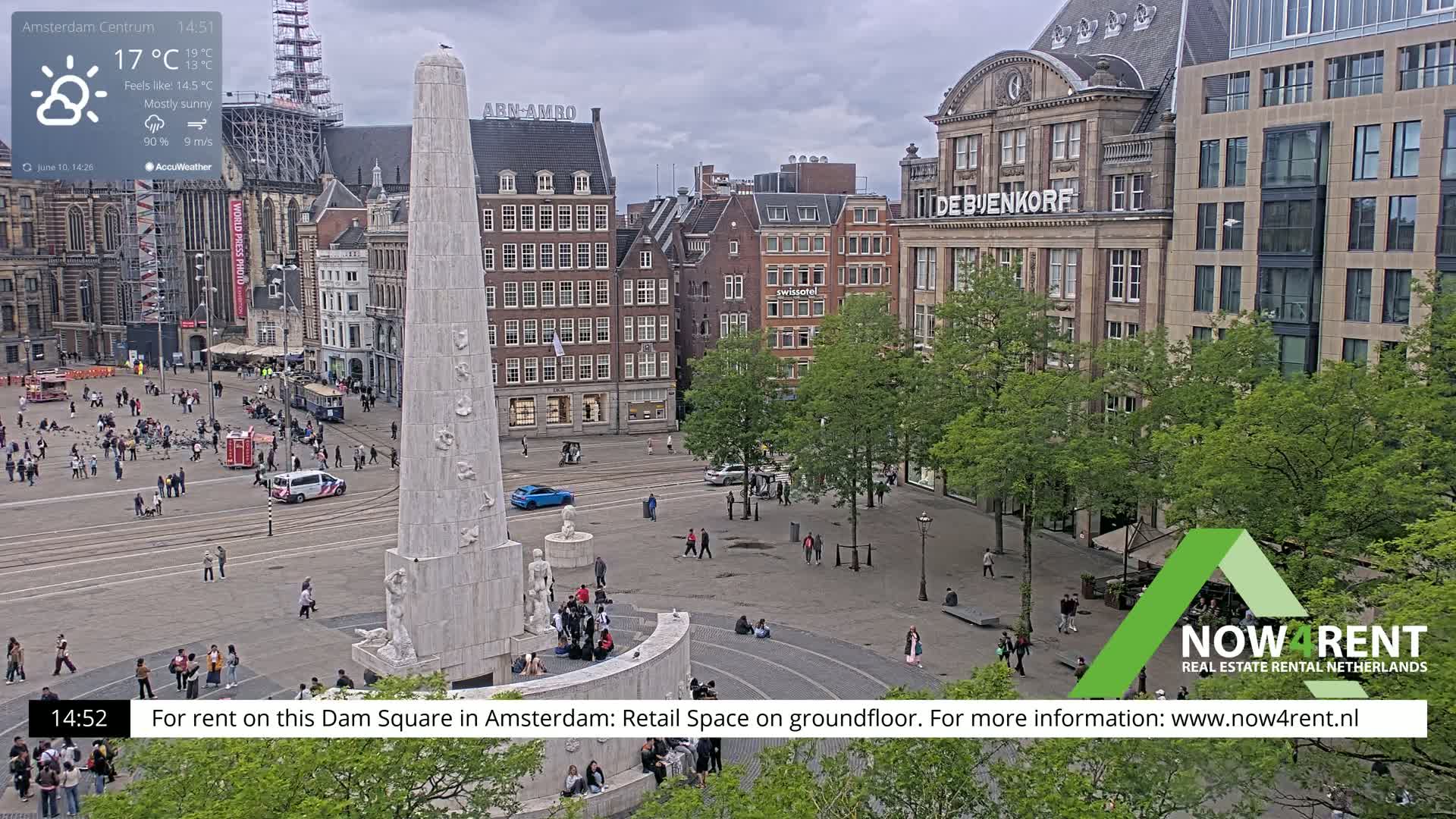 A mostly sunny day reveals a wide, paved square in a European city, featuring a tall obelisk-like monument surrounded by pedestrians and buildings, with some trees providing shade.