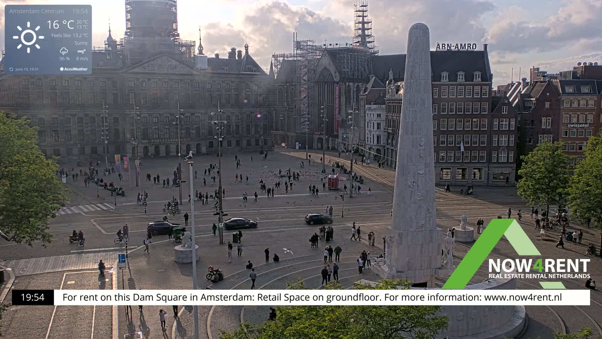 A sunny Dam Square in Amsterdam, with a large monument in the foreground, shows many people and vehicles scattered around the square, with buildings surrounding it under a partly cloudy sky.