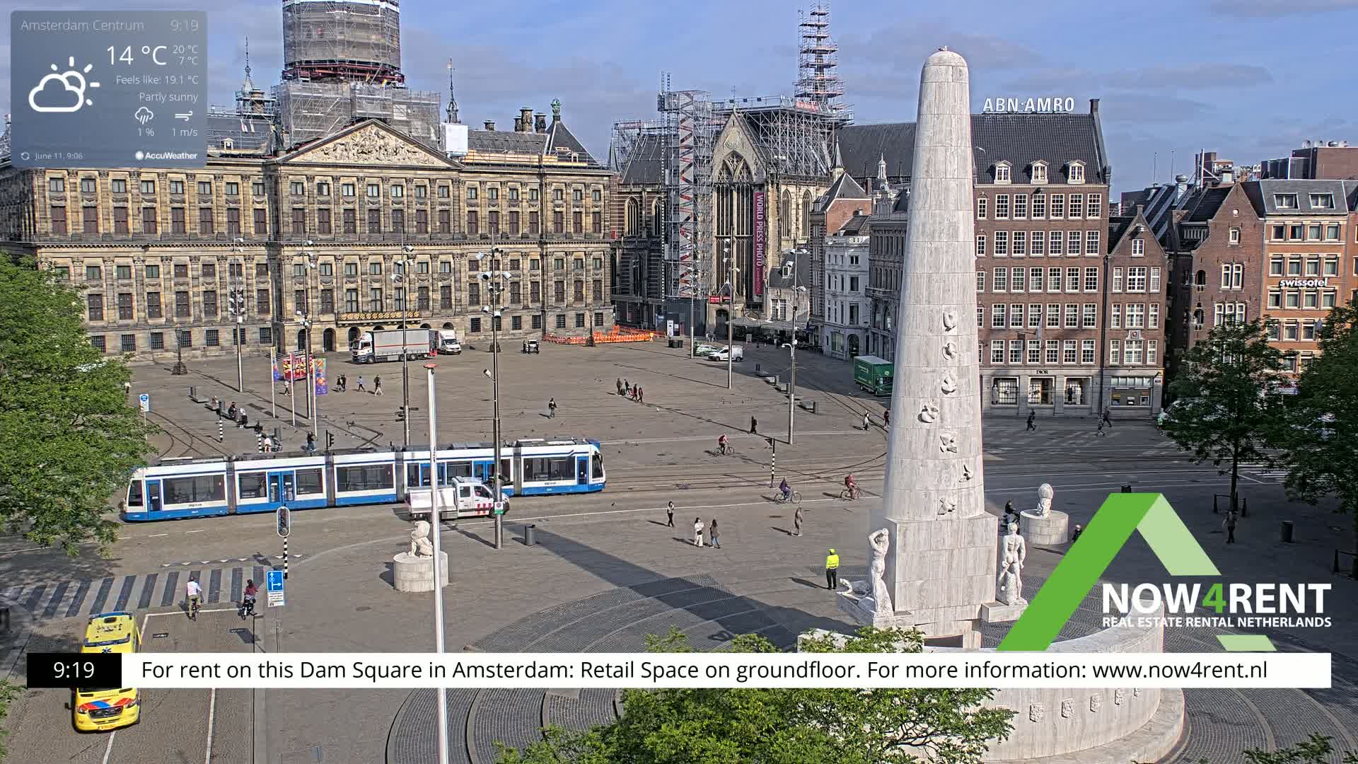 A partly sunny city square with a tram, monument, and large buildings, under a clear sky.
