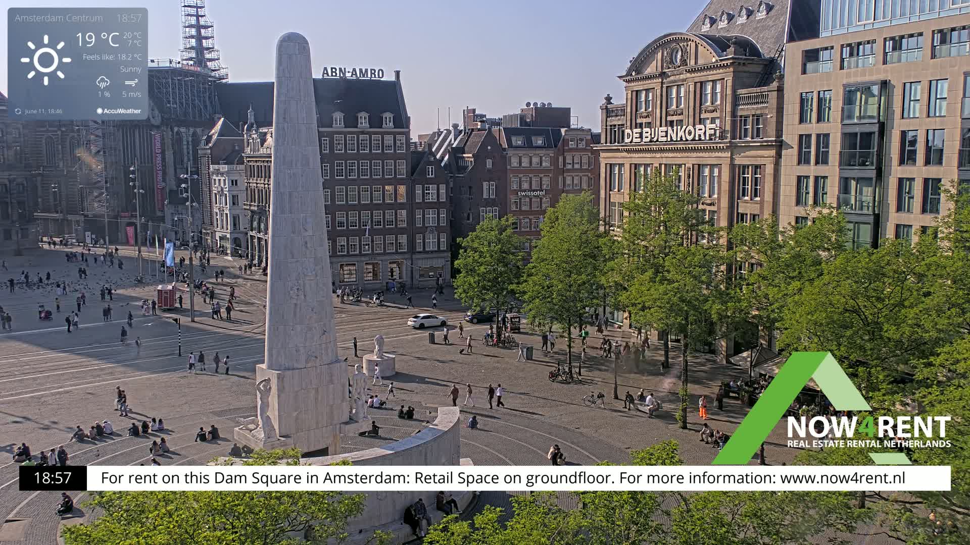A sunny view of Dam Square in Amsterdam, showing a monument, surrounding buildings, and people walking and sitting in the square.