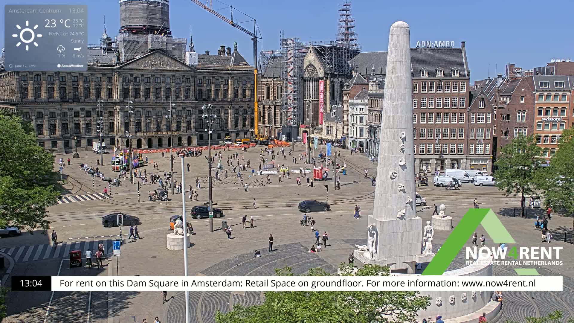 A sunny day in a large European city square, showing a monument, several buildings, and many people walking around.
