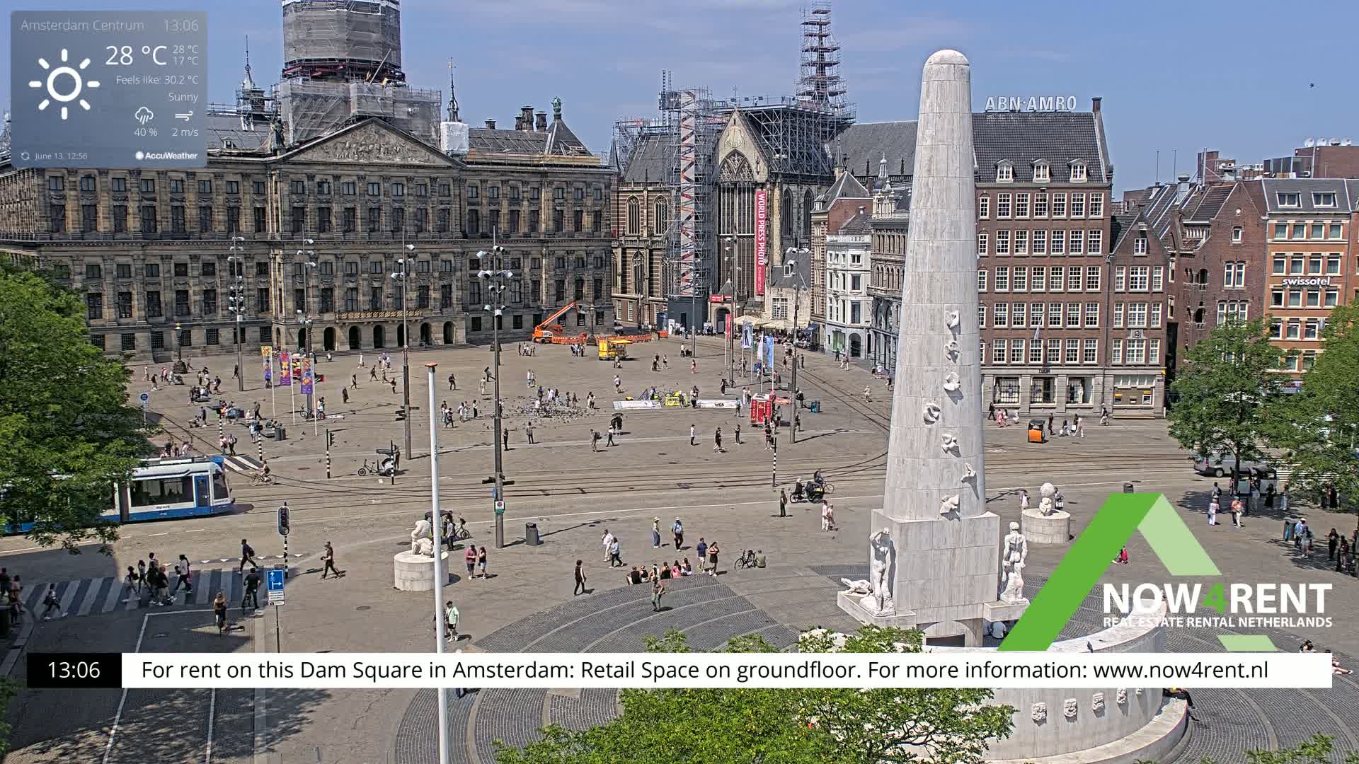 A sunny Dam Square in Amsterdam, showing a monument, surrounding buildings, and people, with some construction visible.