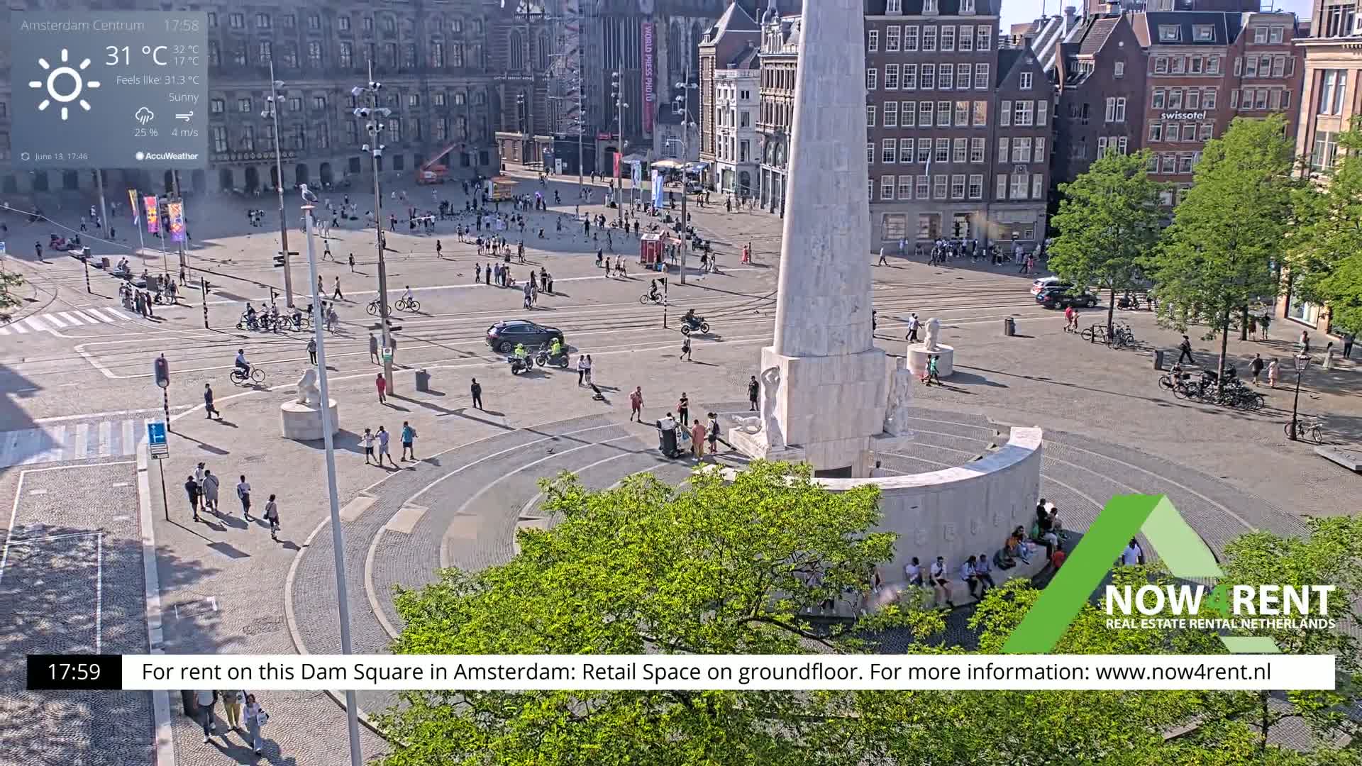 A sunny Dam Square in Amsterdam shows a monument surrounded by people and vehicles, with some cyclists and pedestrians on the surrounding streets.