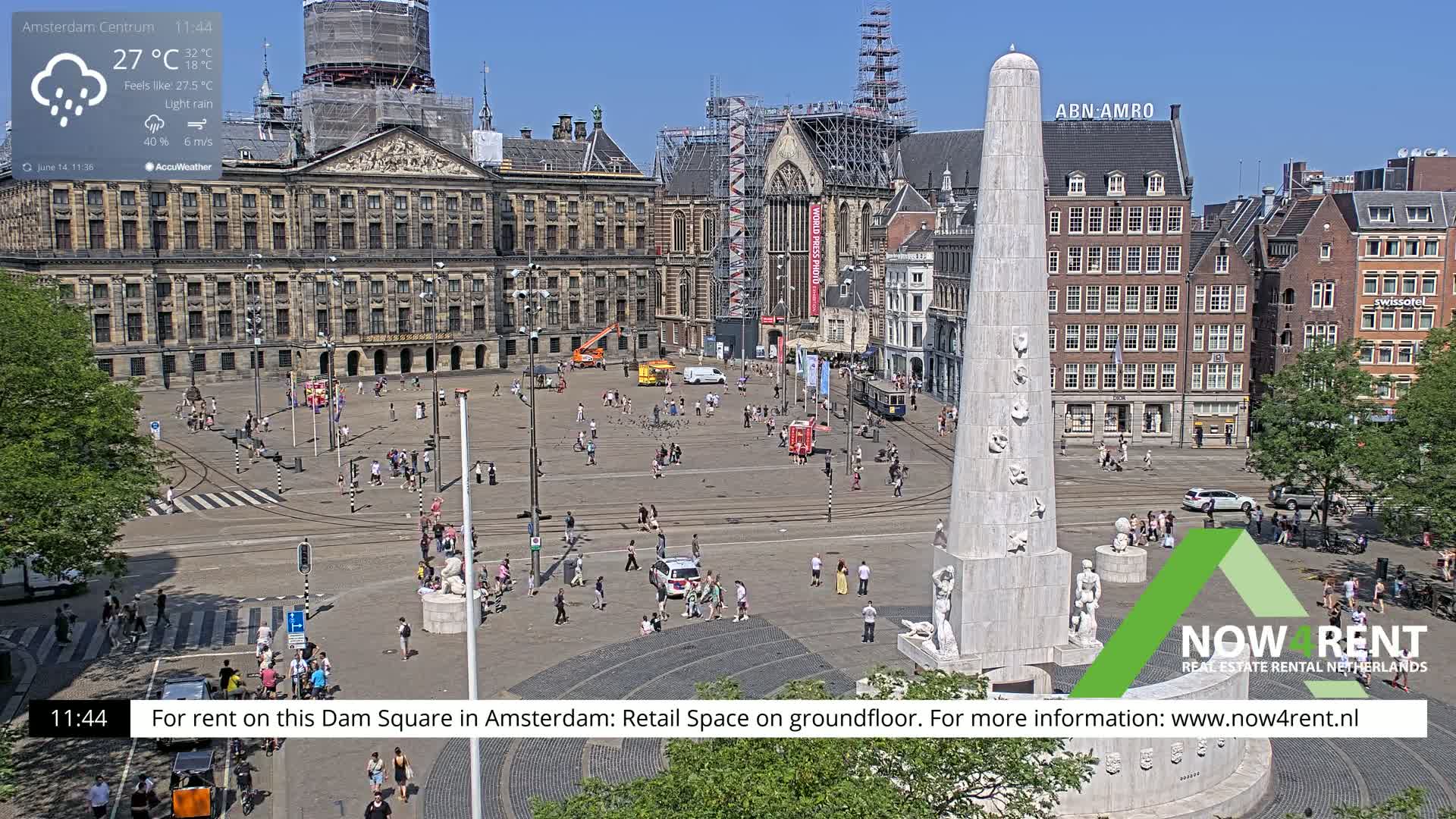 A large, bustling town square with a prominent monument at its center, under light rain on a sunny day.