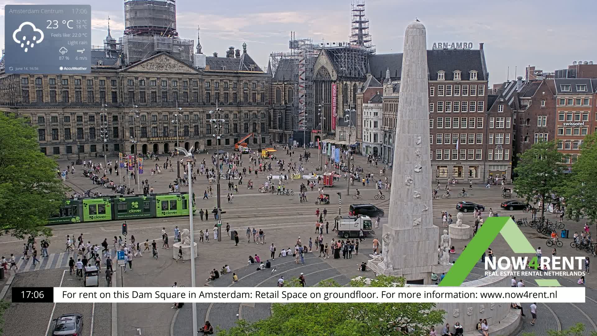 A bustling city square with a tall monument at its center, under light rain and overcast skies, features a tram and numerous people walking and sitting around the square, with several buildings surrounding it, one of which is undergoing construction.