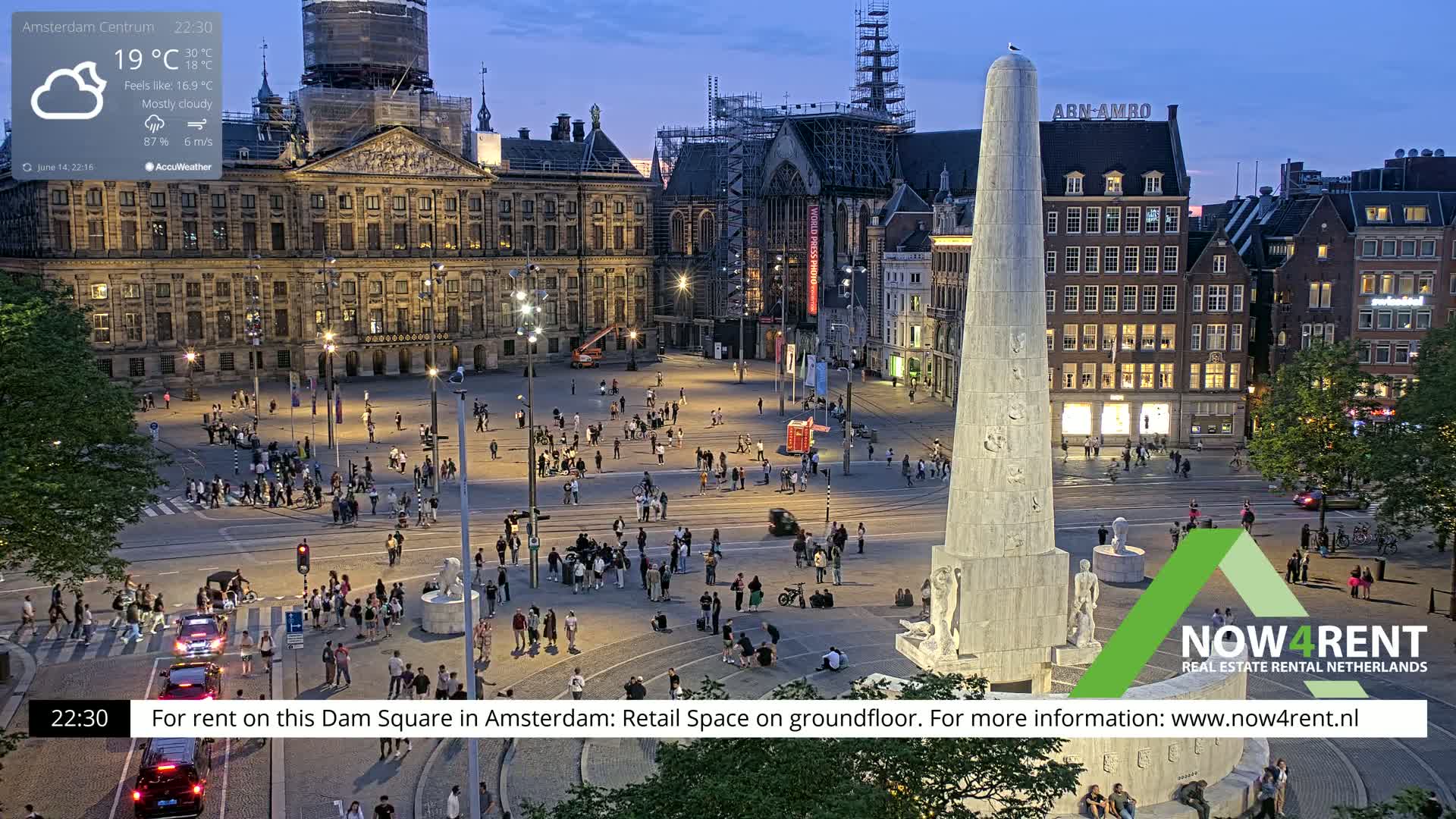 A bustling city square with a tall monument at its center, under light rain and overcast skies, features a tram and numerous people walking and sitting around the square, with several buildings surrounding it, one of which is undergoing construction.