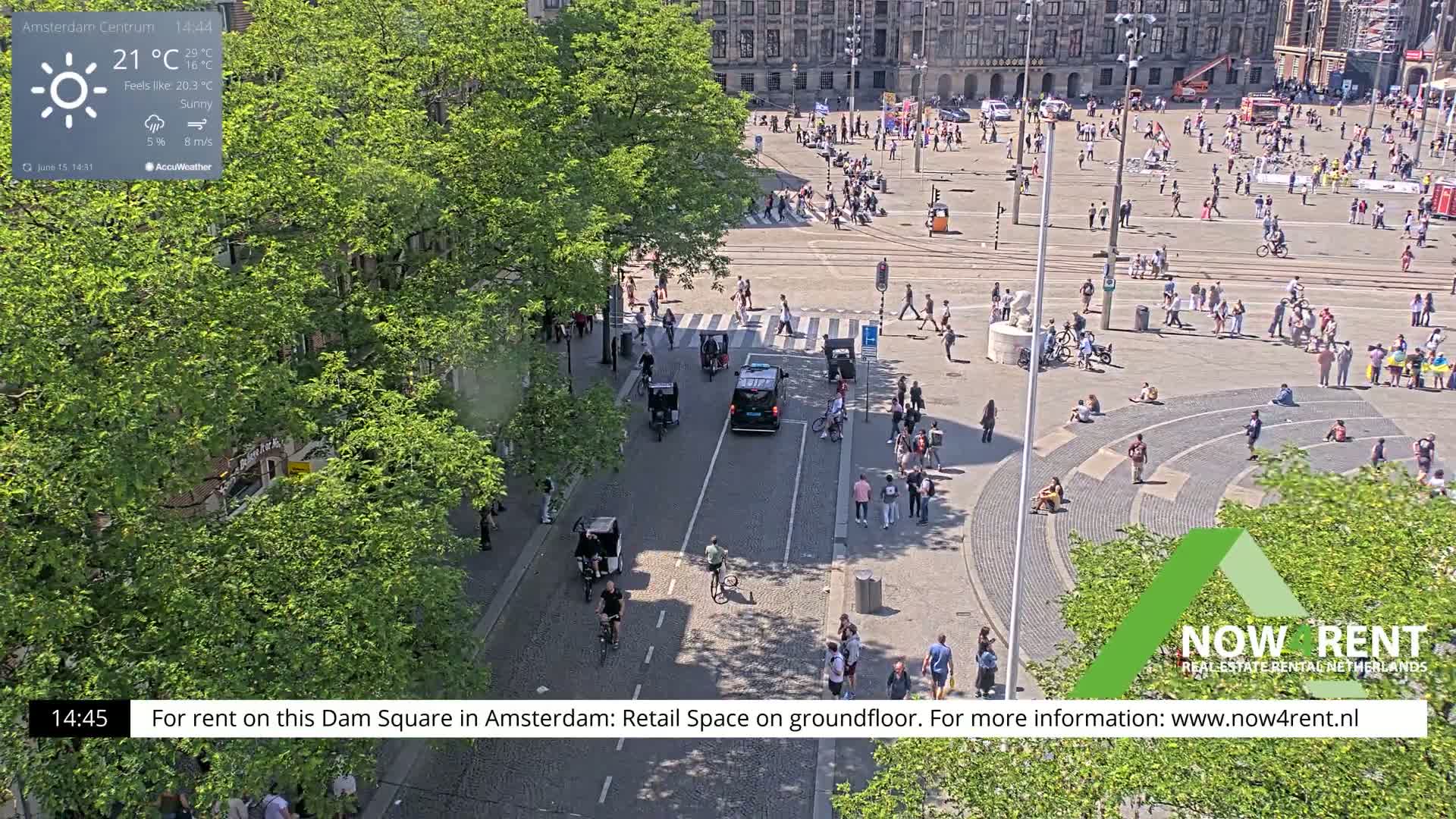A sunny Dam Square in Amsterdam, with numerous people walking and sitting, is viewed from above, showing a street with cyclists and vehicles.