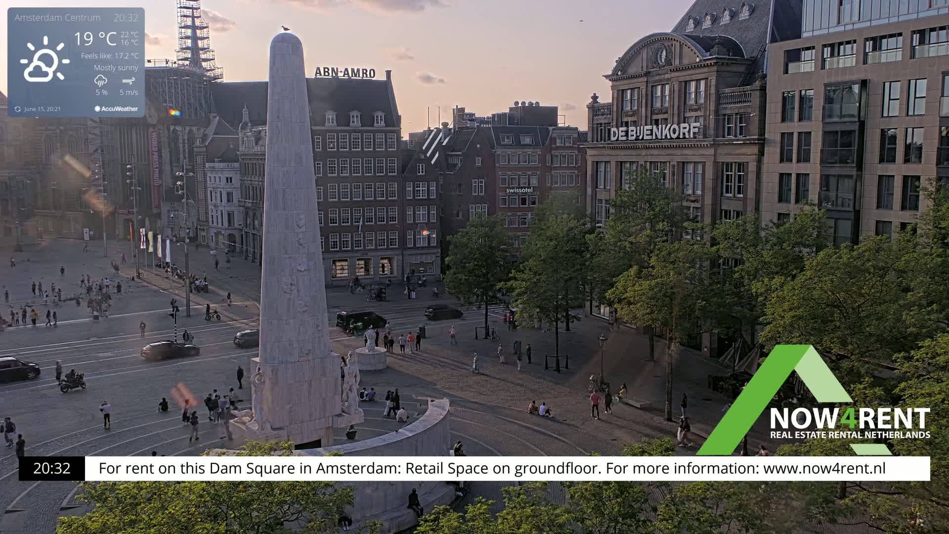 A mostly sunny, late evening view of Dam Square in Amsterdam, showing a monument, surrounding buildings, and people walking around.