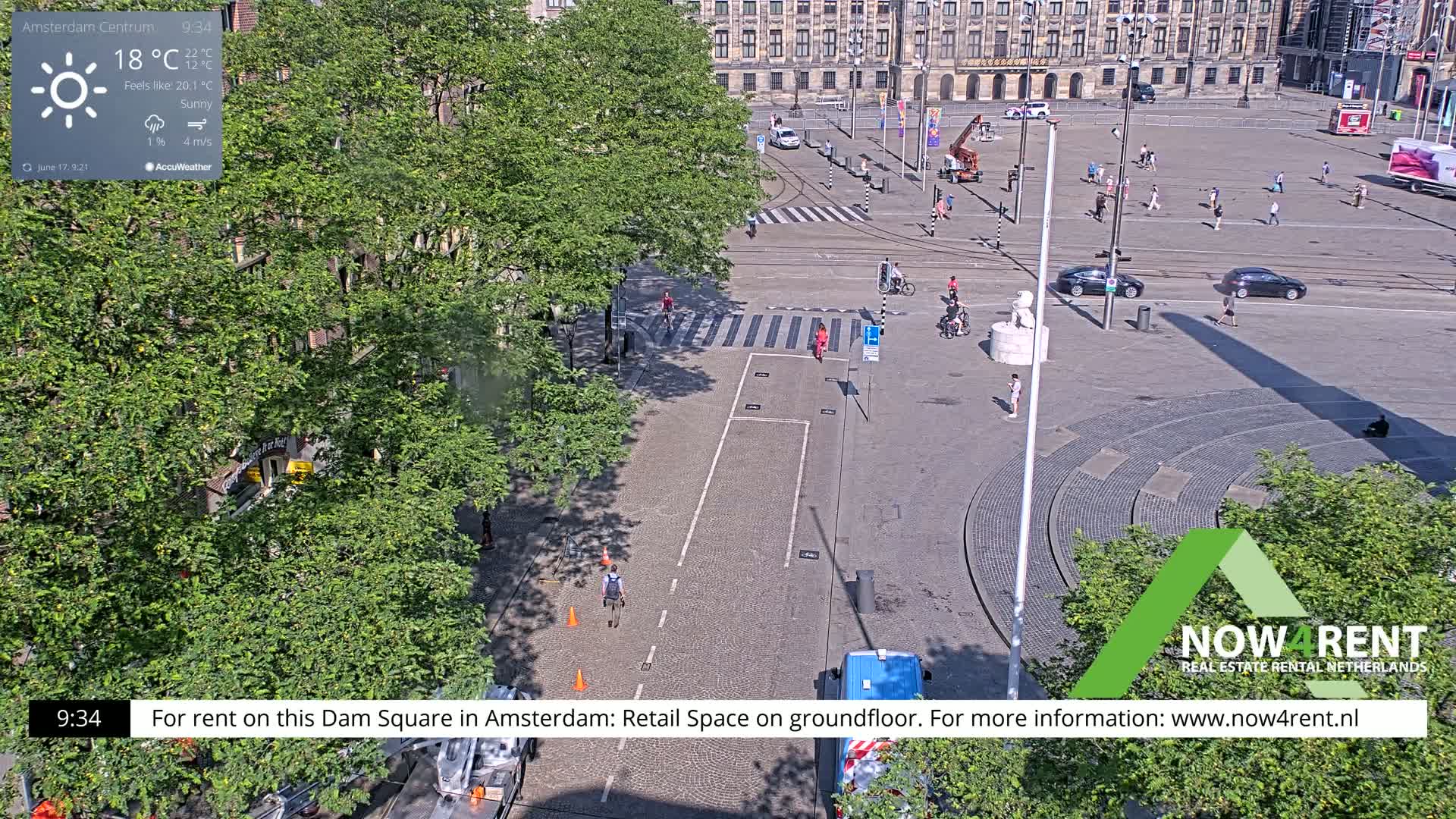 A sunny Dam Square in Amsterdam shows a few people walking and biking on a paved area, with cars parked nearby and buildings in the background.