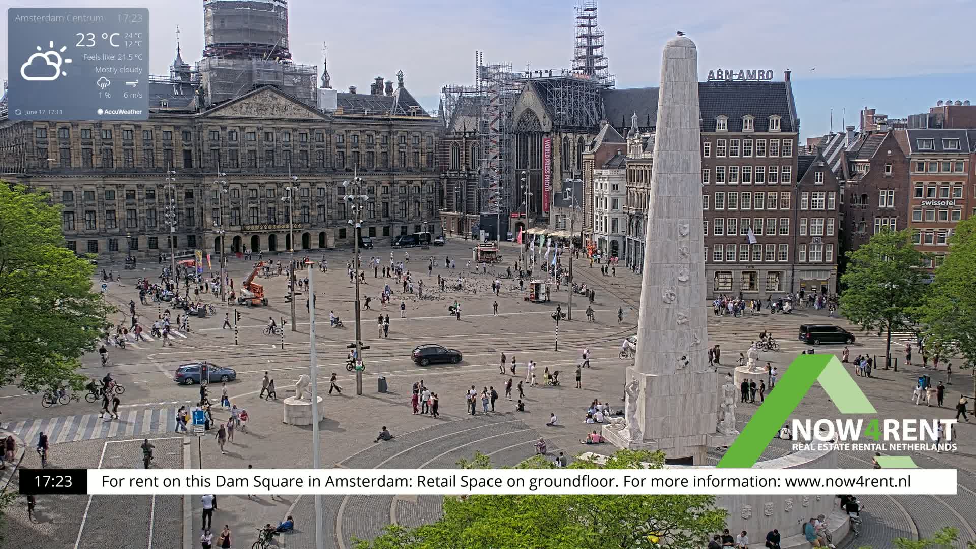 A mostly cloudy, daytime view of Dam Square in Amsterdam shows a large crowd of people gathered around a tall, light-colored monument, with buildings and scaffolding visible in the background.