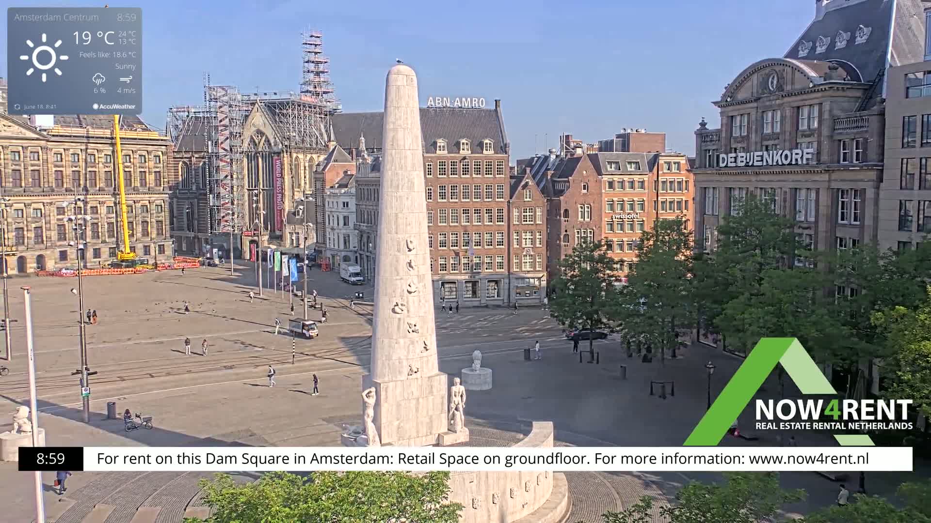 A sunny Amsterdam square, centered around a tall monument, is surrounded by historic buildings under a clear sky.