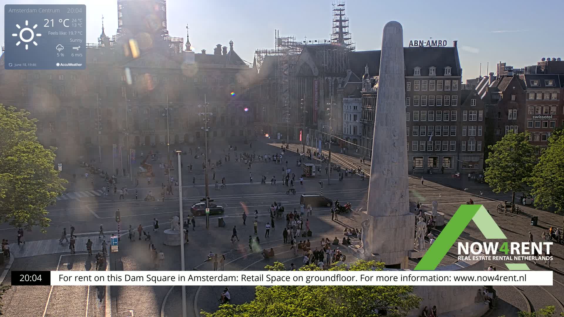 A sunny Dam Square in Amsterdam, with many people gathered around a tall monument, and buildings surrounding the square under a partly sunny sky.