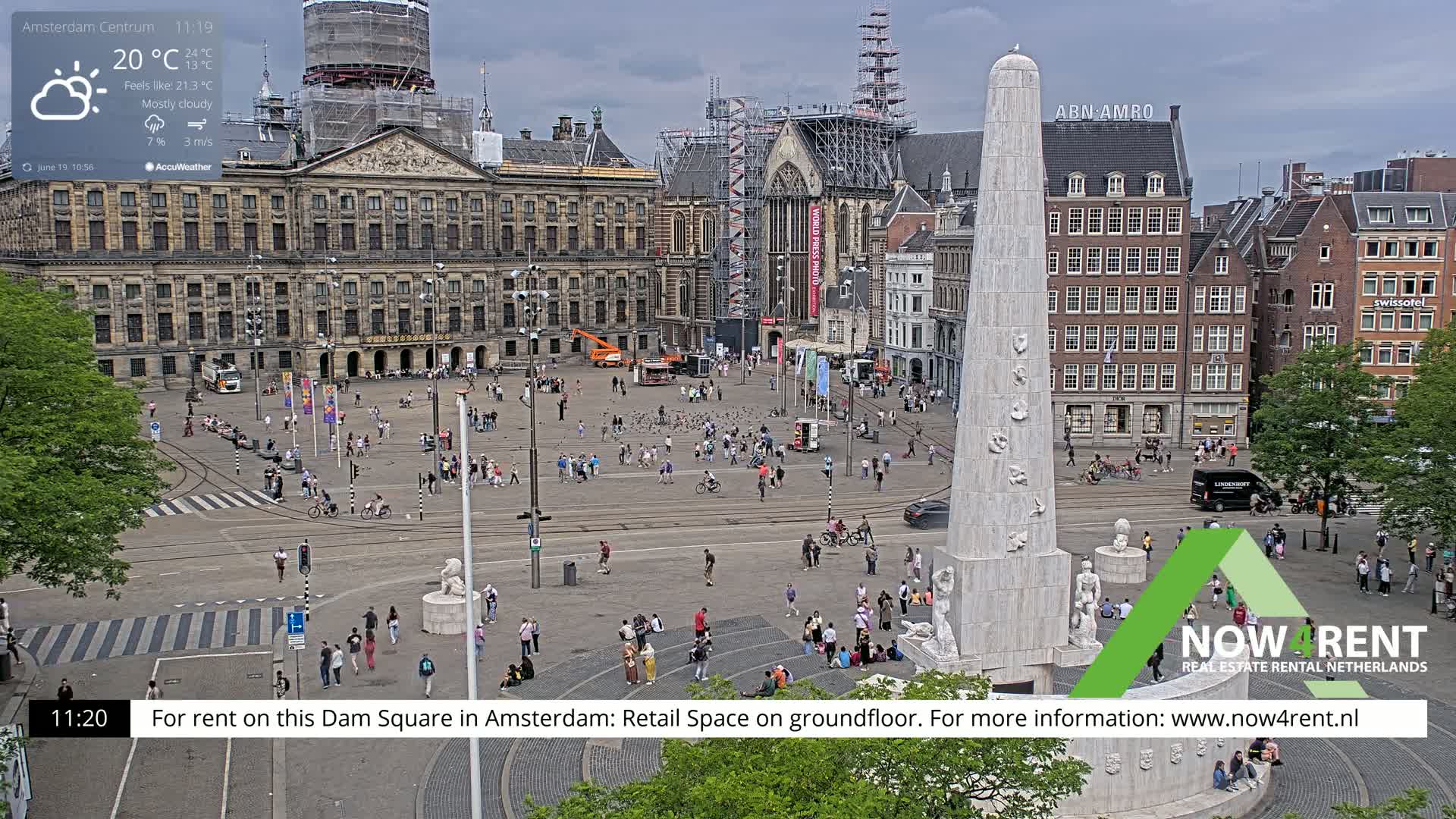 A mostly cloudy day in a large, crowded city square with a tall monument at its center, surrounded by historical buildings, some of which are under construction.