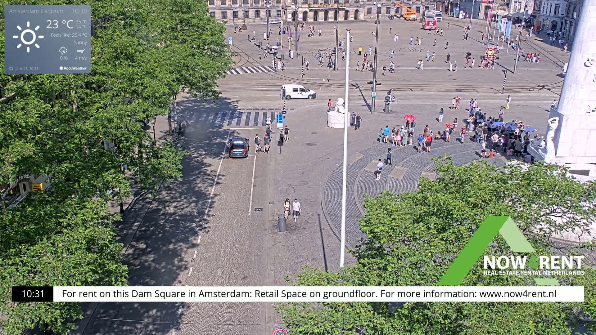 A sunny city square with people walking and gathering around a monument, viewed from above.