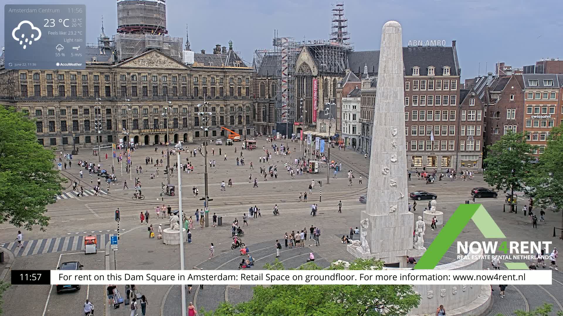 A large square in a city, under light rain, features a tall monument, surrounding buildings, and many people walking around.