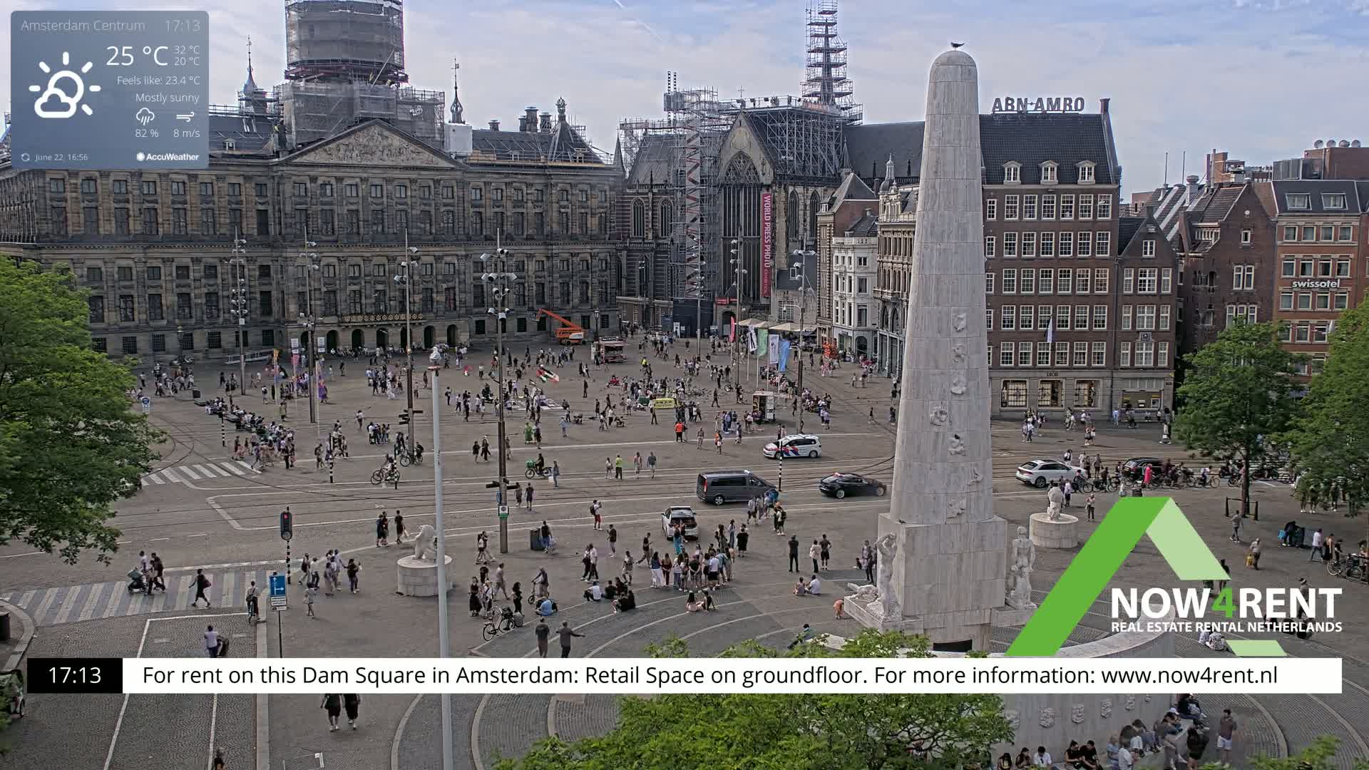 A mostly sunny, crowded city square with a large monument and several buildings, one of which is under construction.
