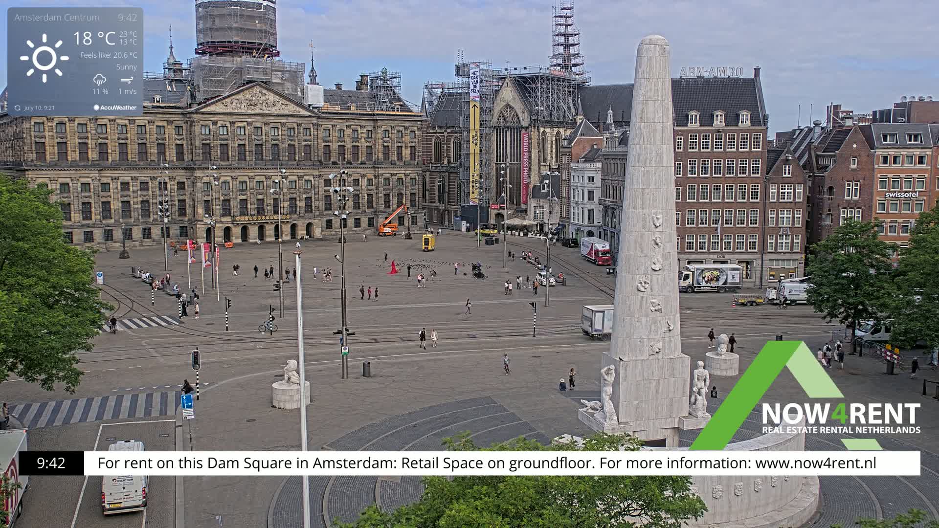 A sunny Dam Square in Amsterdam, with a large monument at the bottom and the Royal Palace in the background, shows a sparsely populated public space with some streetcars and pedestrians.
