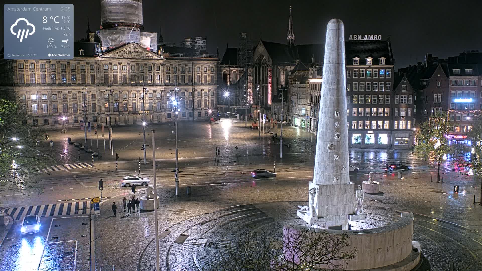 A rainy night view captures a European city square, featuring a prominent obelisk-like monument in the foreground surrounded by illuminated historic buildings and a church under construction.