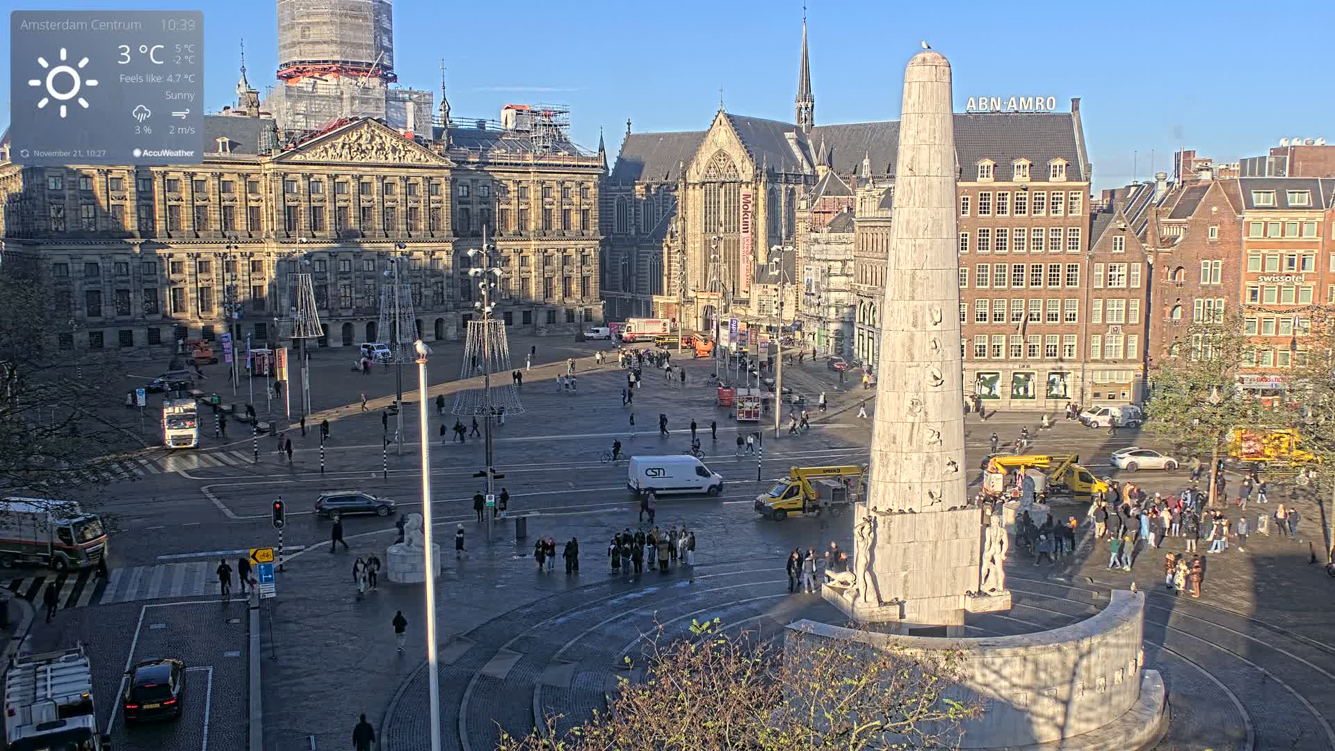A bustling city square is captured on a sunny day, featuring a large classical building, a gothic-style church, a prominent white obelisk monument, and numerous pedestrians and vehicles moving through the space.