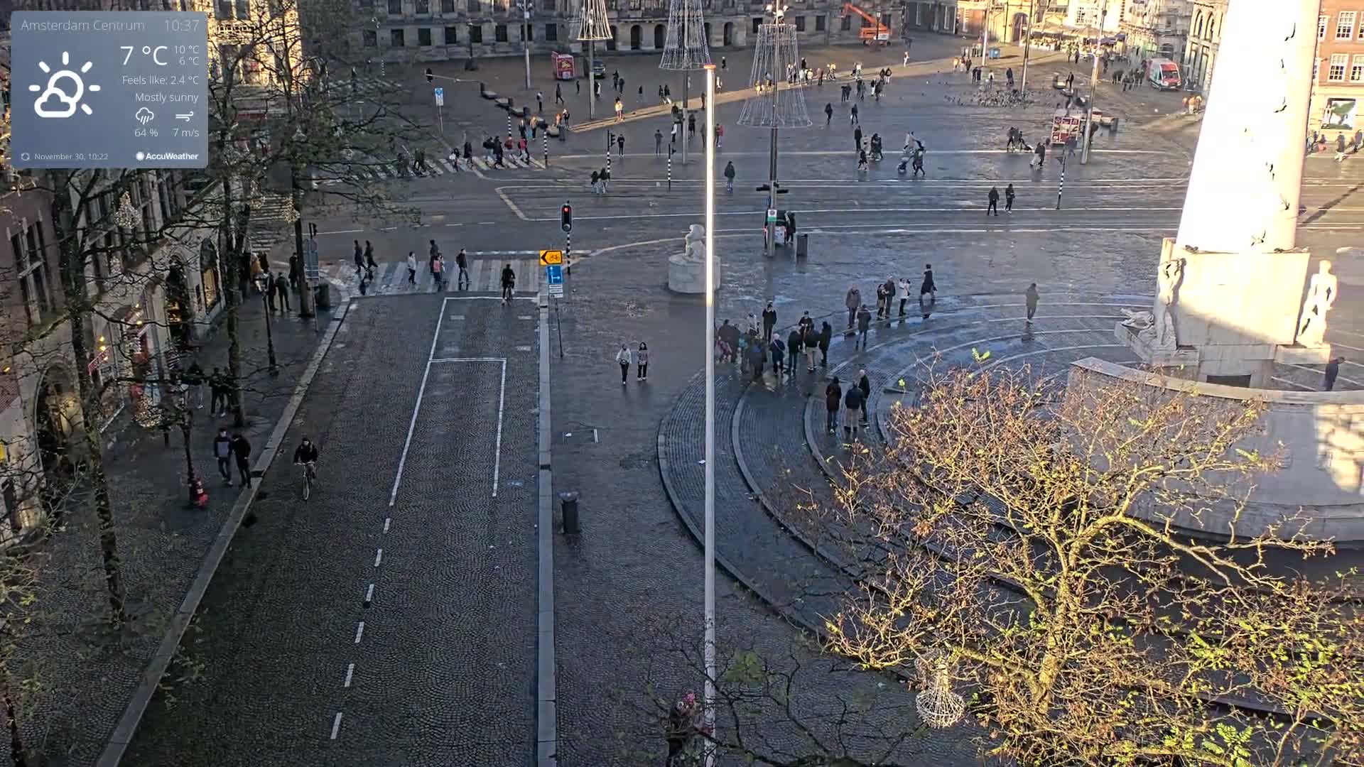 An overhead view reveals a bustling city square with numerous pedestrians, a large stone monument, bare trees, and historic buildings under a cool, partly sunny sky with damp cobblestone streets.