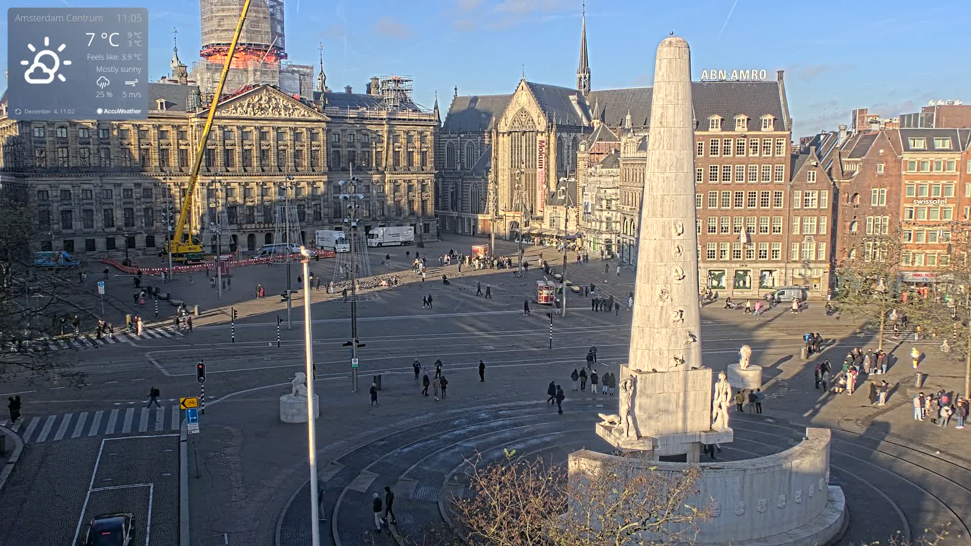 A sunny daytime scene captures a bustling European public square featuring a prominent tall monument, surrounding historic buildings including one partially obscured by a construction crane, numerous pedestrians, and festive light structures being assembled.