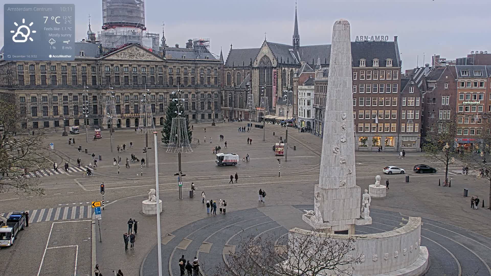 A sunny daytime scene captures a bustling European public square featuring a prominent tall monument, surrounding historic buildings including one partially obscured by a construction crane, numerous pedestrians, and festive light structures being assembled.