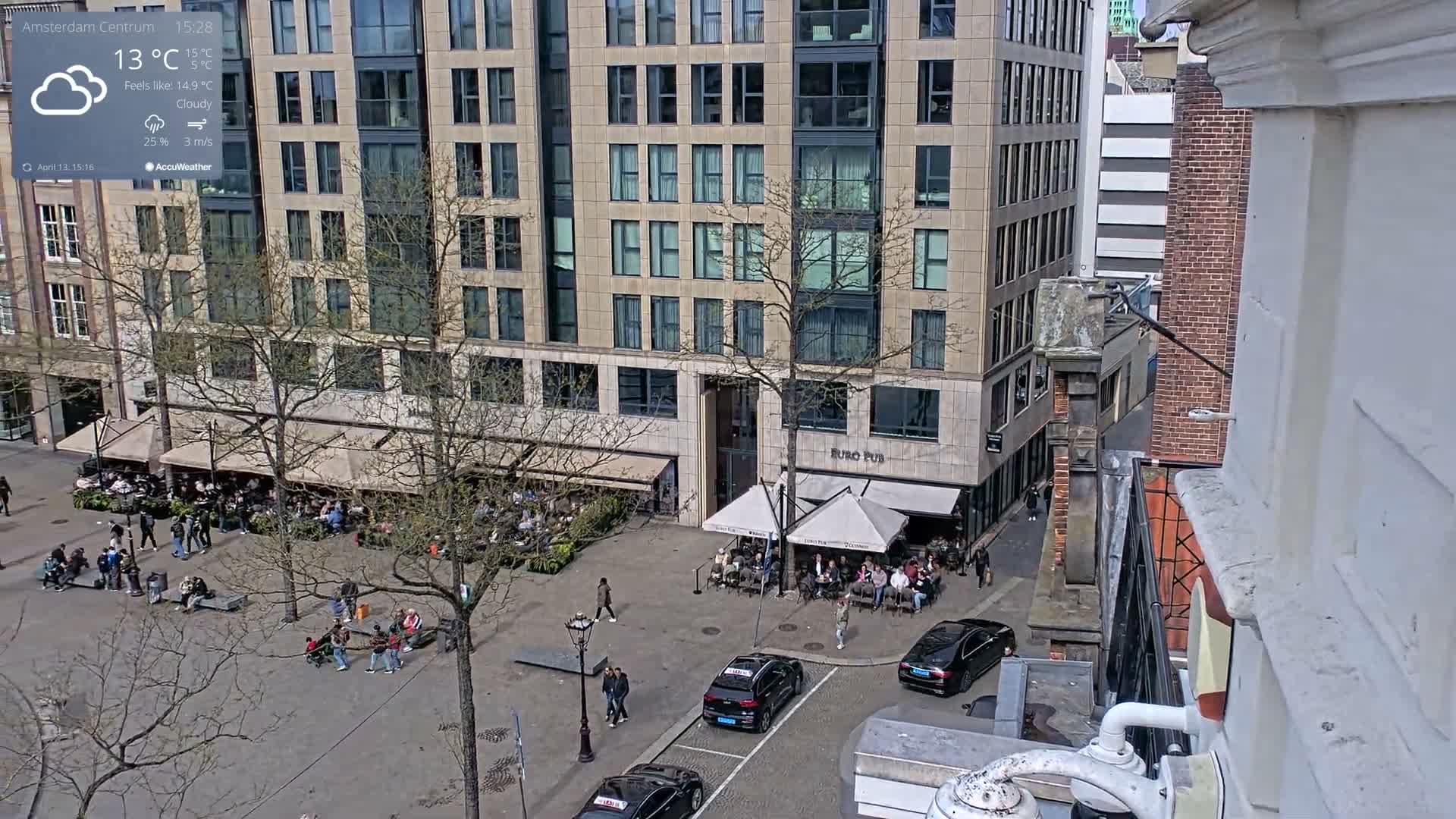 A sunny daytime scene captures a bustling European public square featuring a prominent tall monument, surrounding historic buildings including one partially obscured by a construction crane, numerous pedestrians, and festive light structures being assembled.