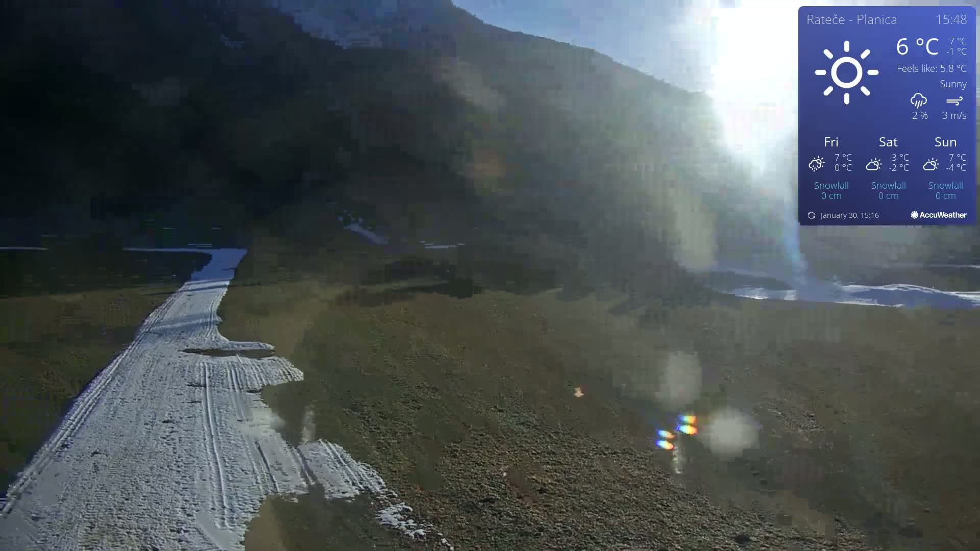 A mostly sunny, snow-dusted landscape shows a partially snow-covered path leading toward a dark mountain under a bright sky.