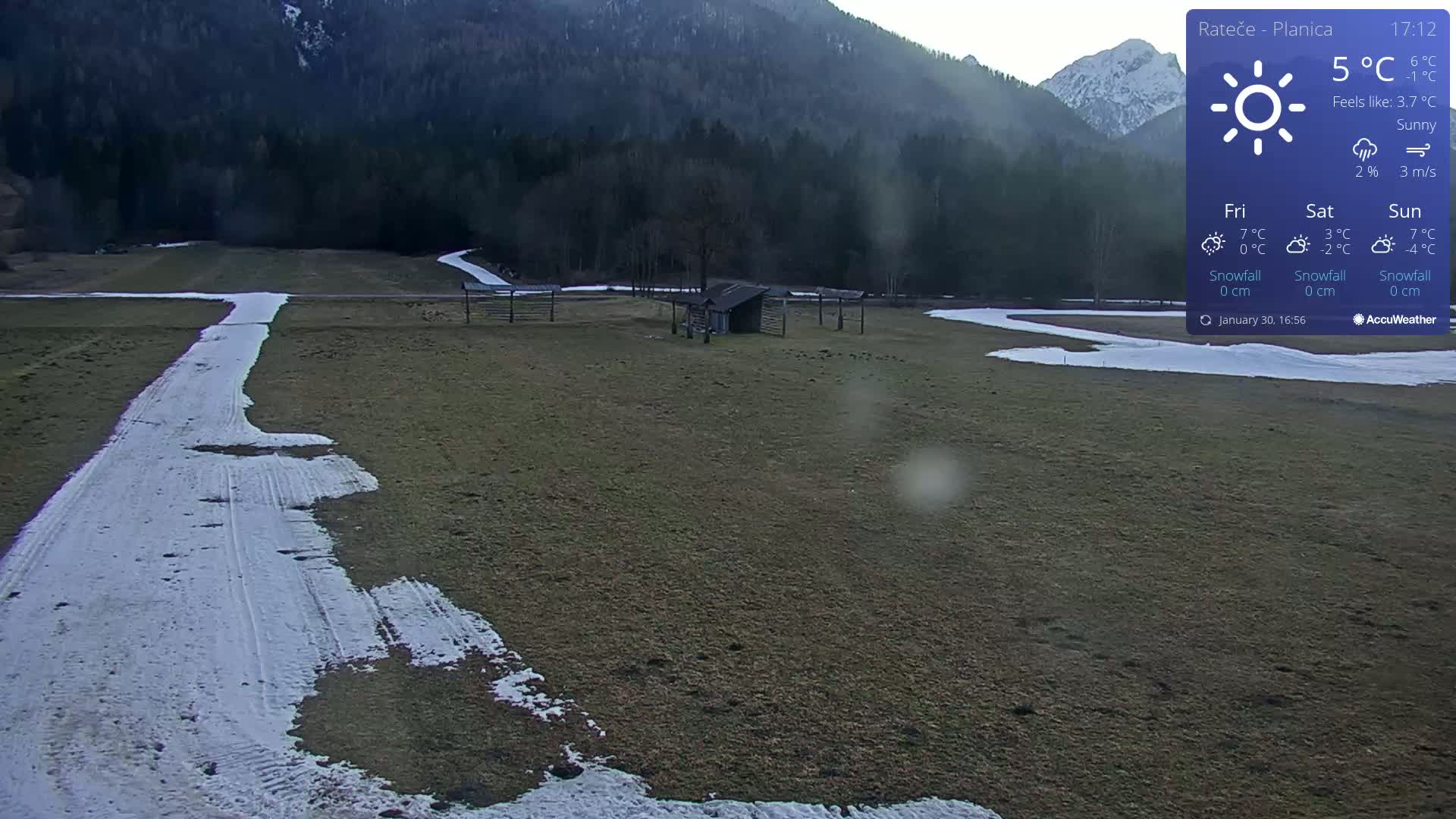 A snowy path winds through a grassy field, past a small shed, towards a backdrop of a mountain range under sunny conditions.