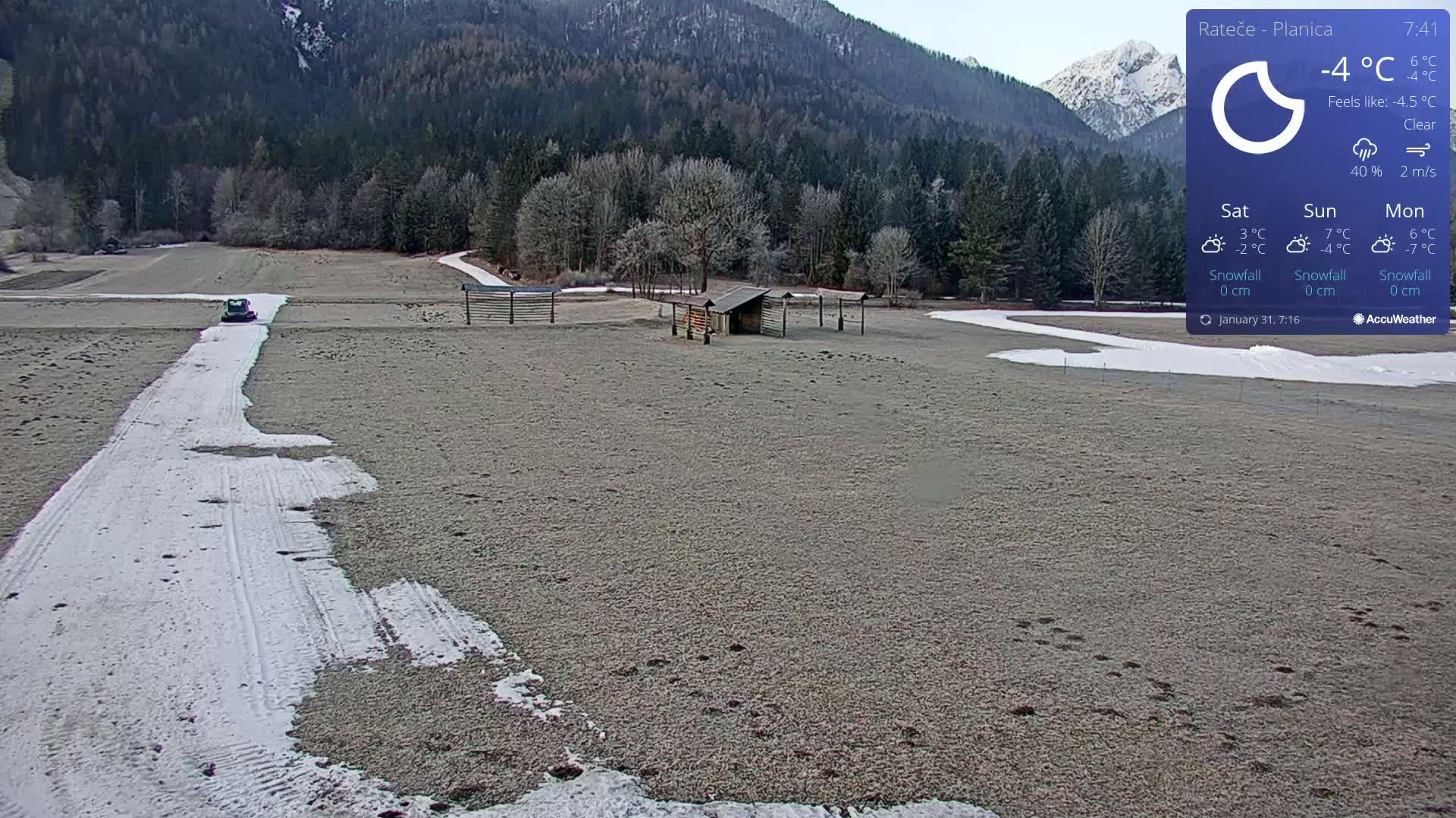 A small vehicle travels down a snow-covered path across a frosty field, with a wooden structure and mountains in the background under clear skies.