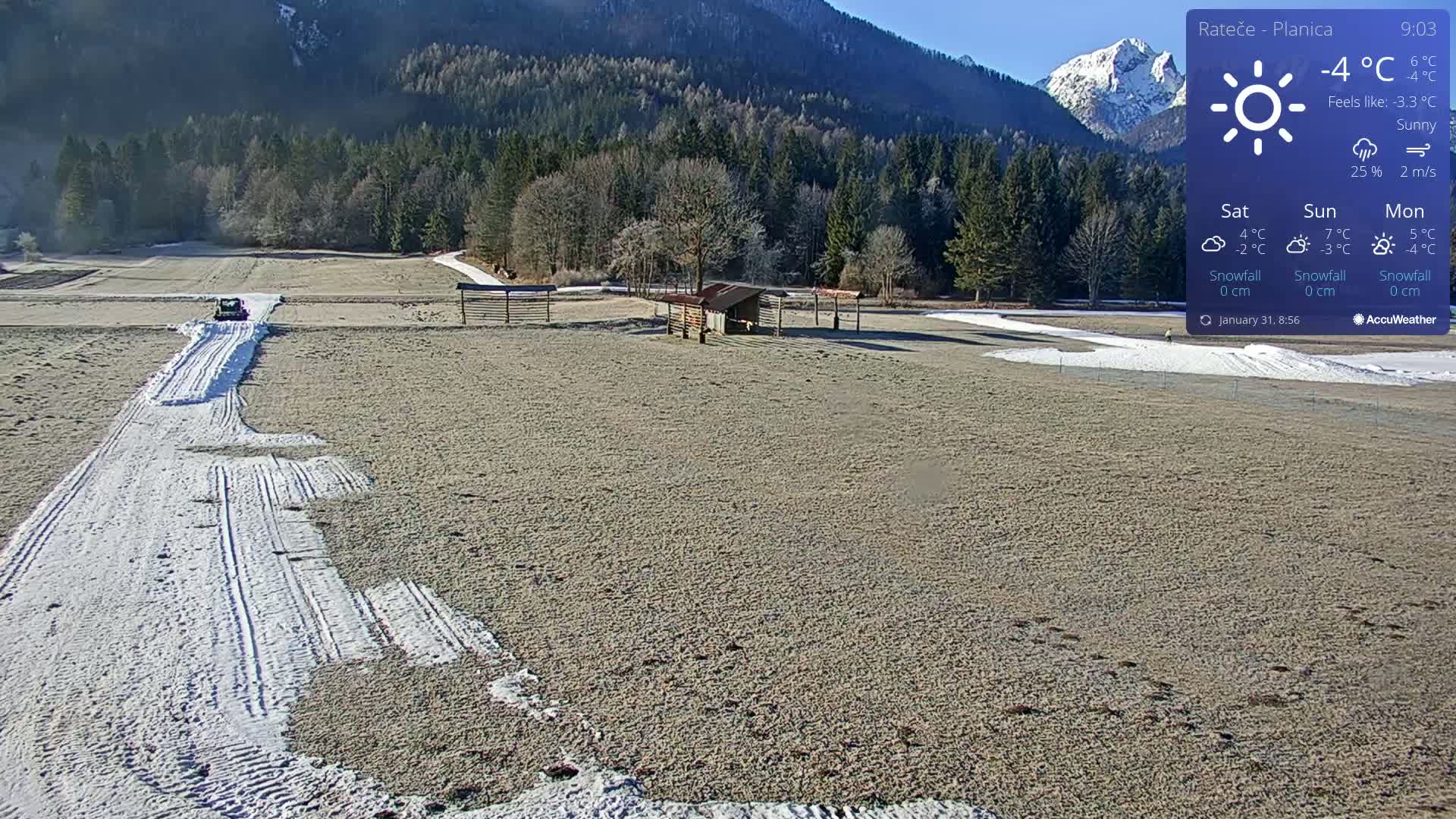 A vehicle is clearing snow from a path across a wide, mostly snow-free field in a sunny mountainous region.