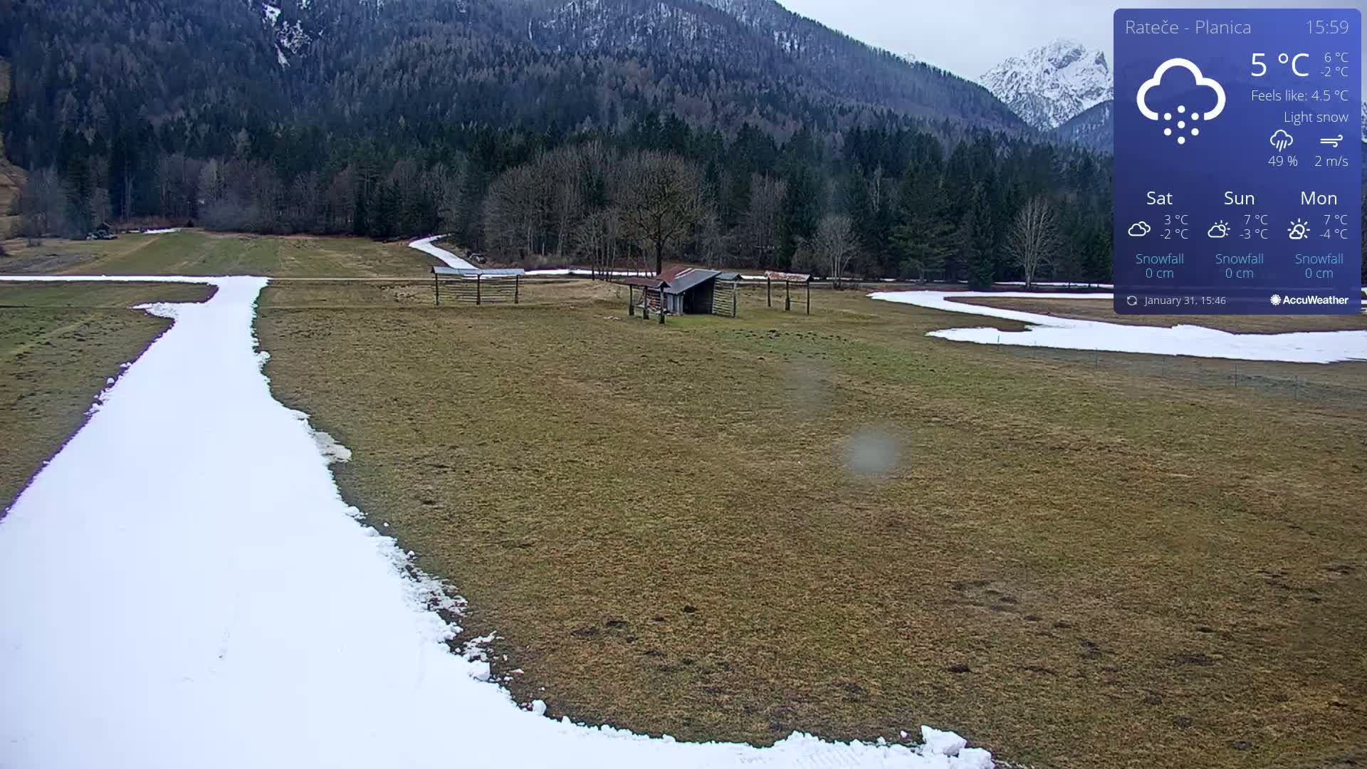 A snow-covered path winds through a grassy field, with a small shed and a backdrop of a mountain range under light snow.