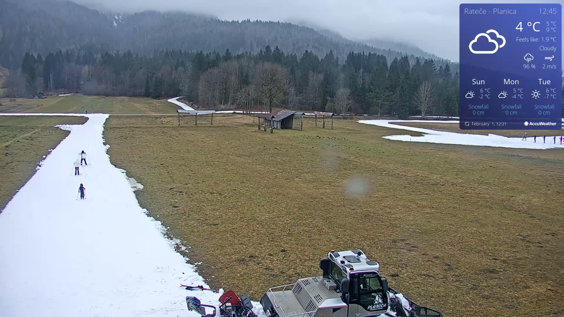 A snowy ski slope with several skiers and a snowcat is visible in a field bordered by a forest under cloudy skies.