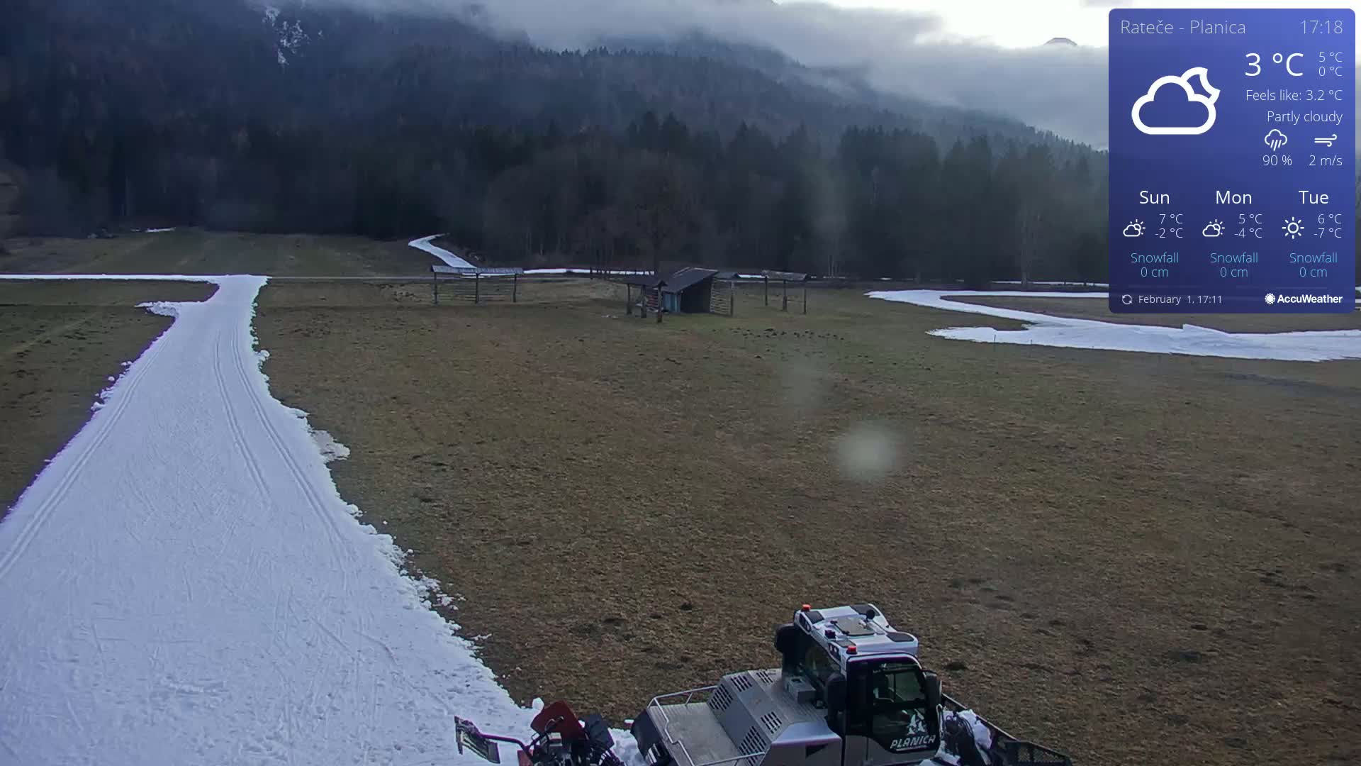A snow groomer is parked on a snow-covered cross-country ski trail in a partly cloudy landscape, with mountains in the background.