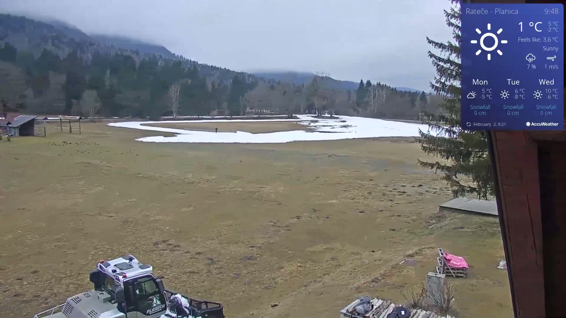 A mostly sunny, cold day reveals a snow-dusted field near a wooded mountain range, with a snowplow visible in the foreground.