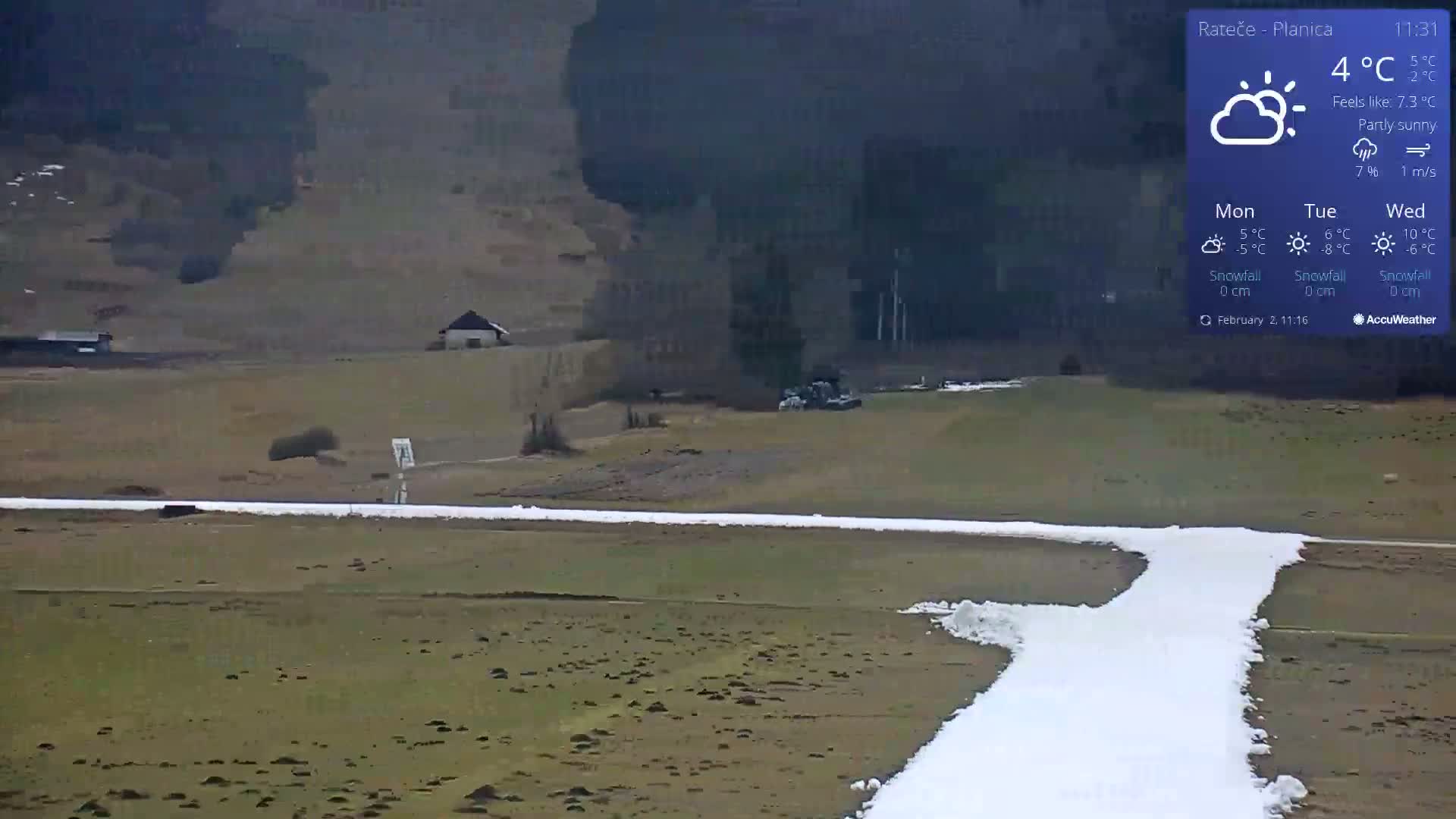 A partly sunny landscape shows a snow-covered path winding through a mostly brown field, with a small building and some machinery visible in the distance.