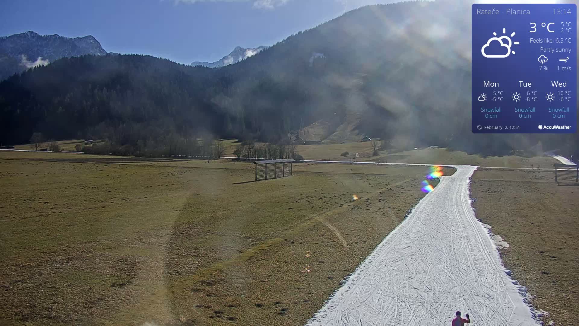 A partly sunny day shows a snow-covered path winding through a field toward snow-capped mountains in the distance.