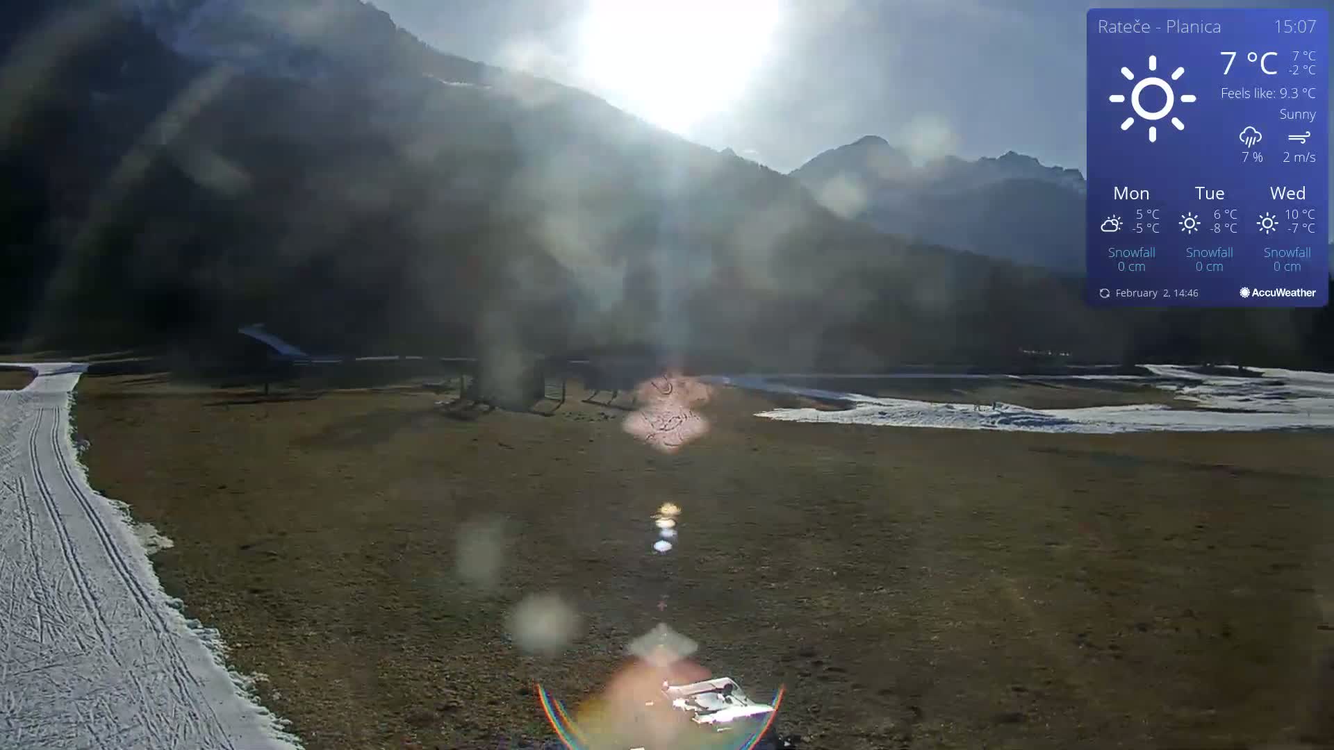 A sunny, snow-dusted field stretches out before a mountain range under a bright sun, with some lens flare visible.