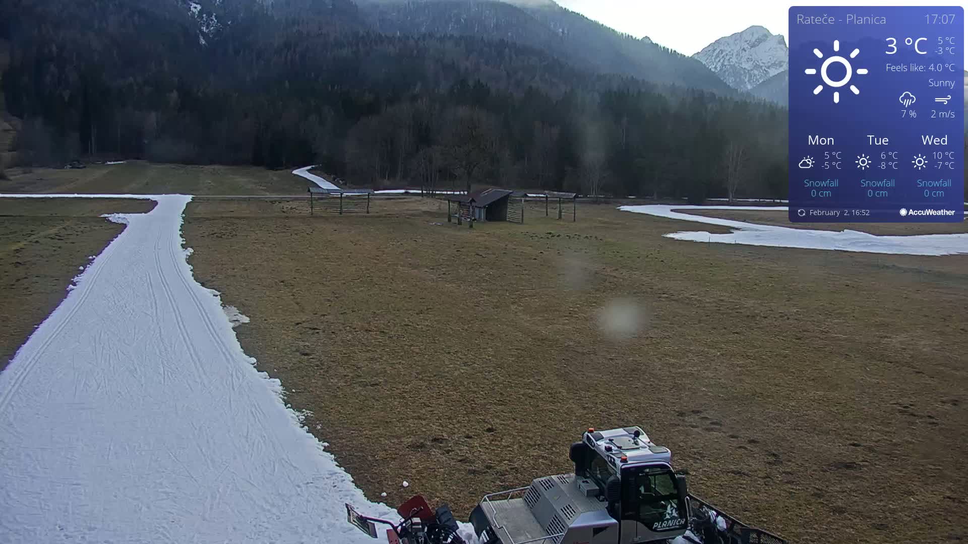 A snow groomer is parked at the end of a groomed ski trail in a sunny, sparsely snow-covered field, with mountains in the background.