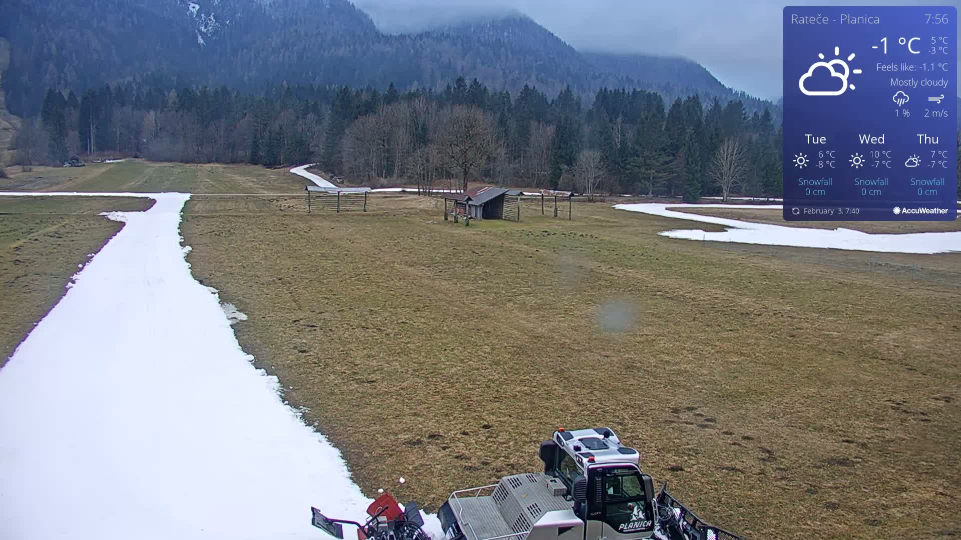 A snow groomer prepares a snow-covered path in a mostly cloudy, -1°C field surrounded by a forest and mountains.