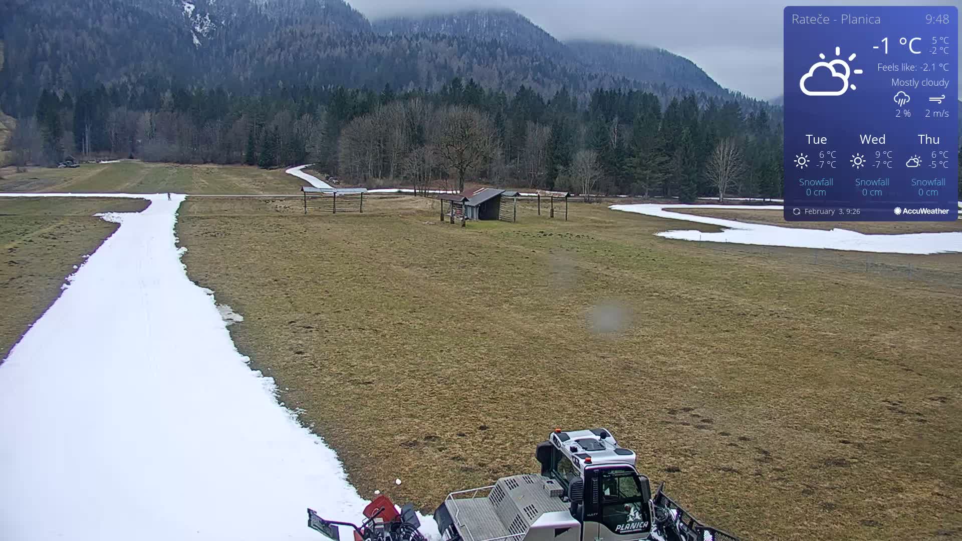 A snow groomer is parked on a mostly brown field with a long, narrow snow path leading toward a line of trees and mountains under mostly cloudy skies.