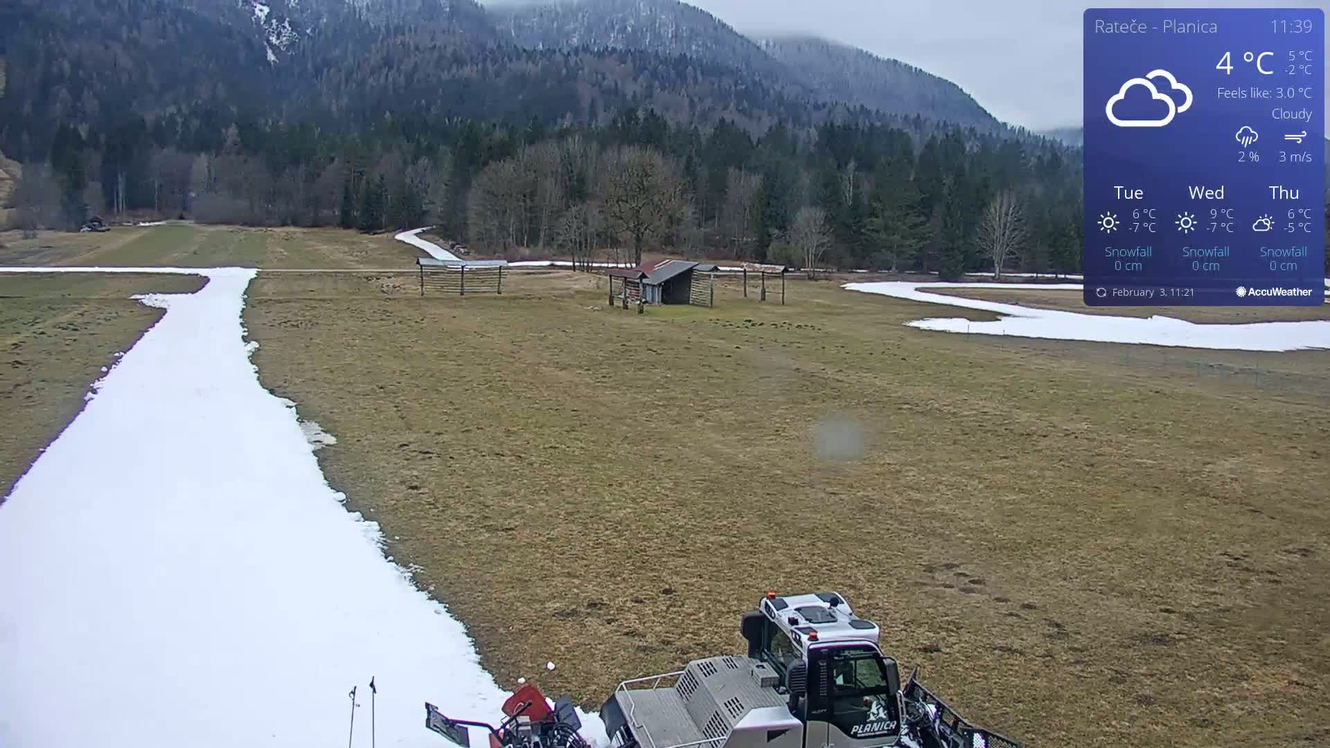 A snow groomer is parked on a grassy field with a snow-covered path, under a cloudy sky.
