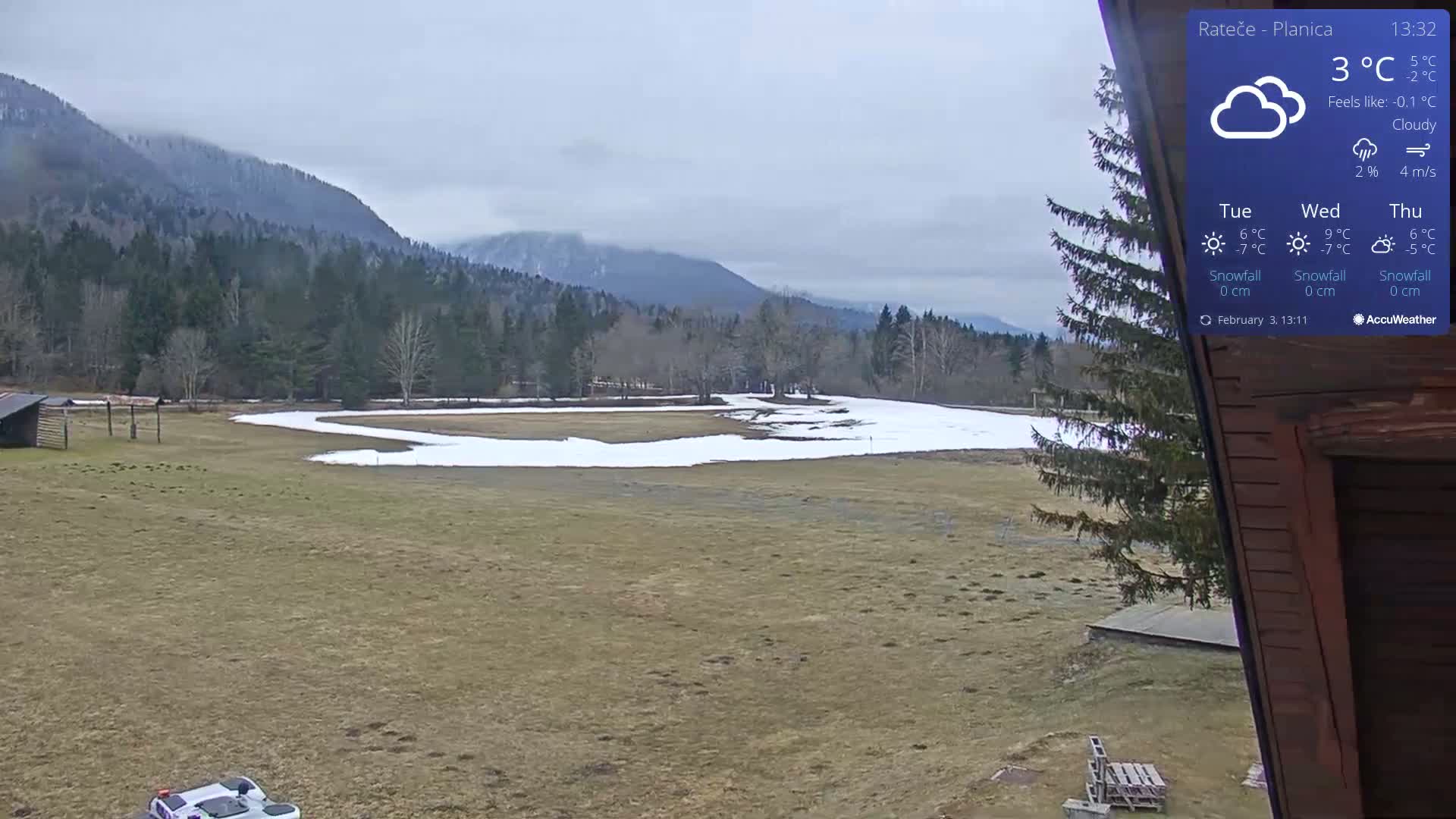 A mostly cloudy view of a snow-dusted field with a small wooden building and distant mountains.