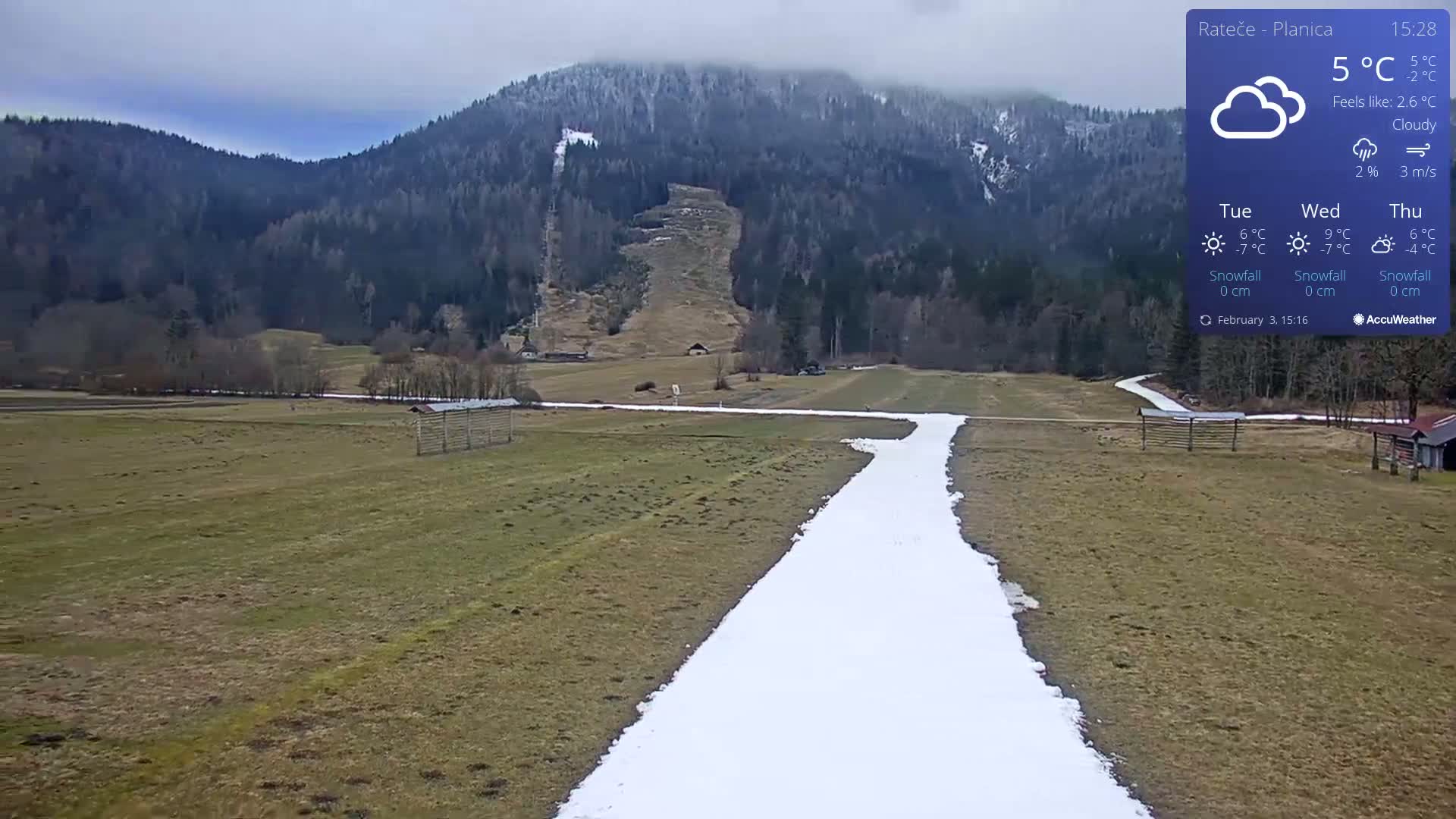 A long, narrow path of snow winds through a grassy field toward a mountainous background under cloudy skies.