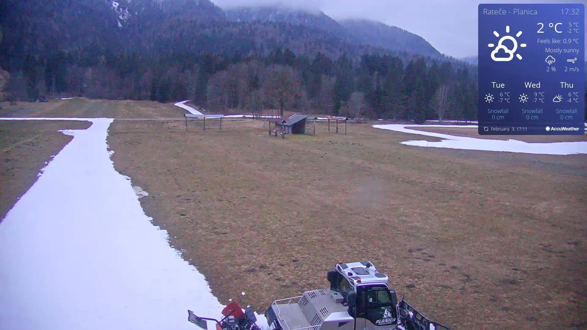 A snowcat grooms a snow-covered path in a mostly sunny, outdoor field bordered by a forest and mountains.