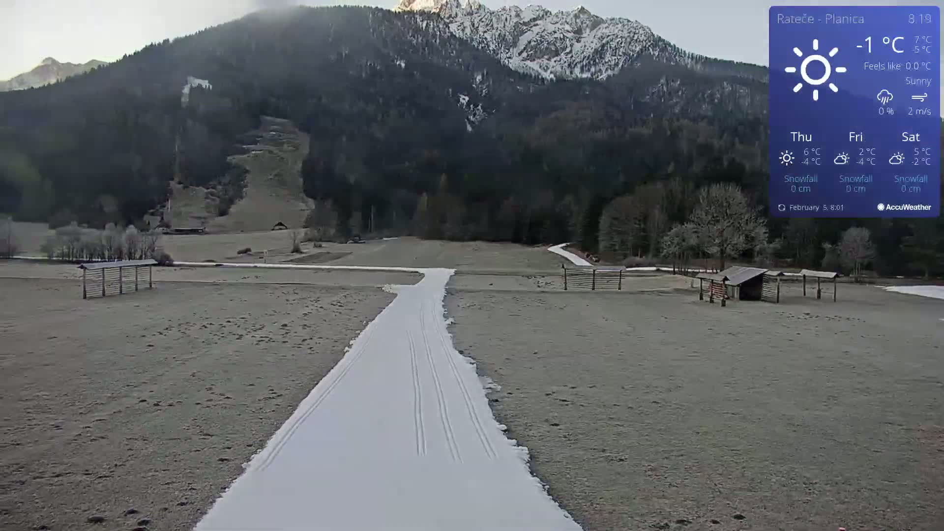 A snowy cross-country ski trail stretches across a frosty field towards a mountainous backdrop under sunny skies.