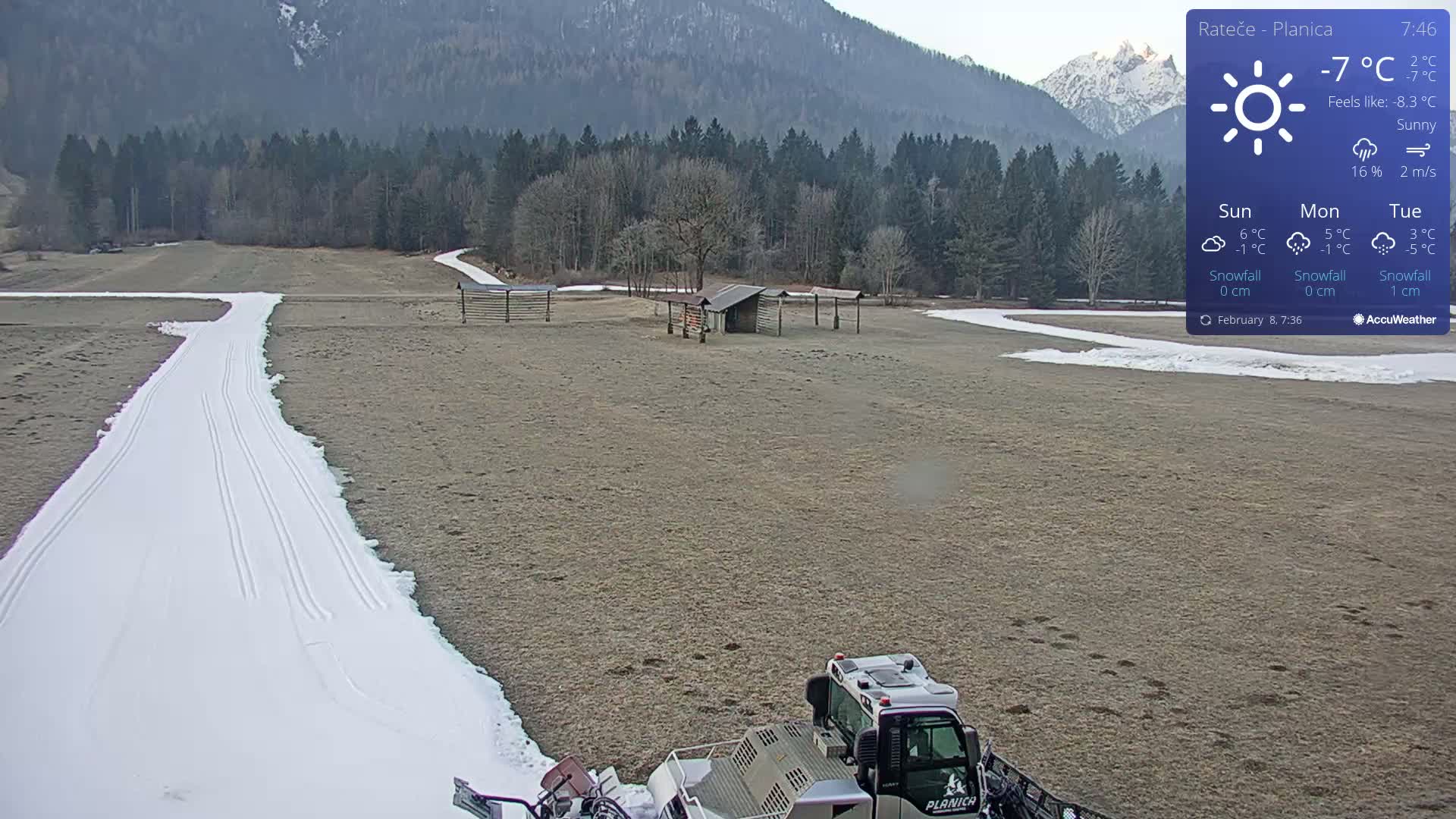 A snow groomer is preparing a snow-covered cross-country ski trail in a sunny, cold field against a backdrop of mountains and a coniferous forest.