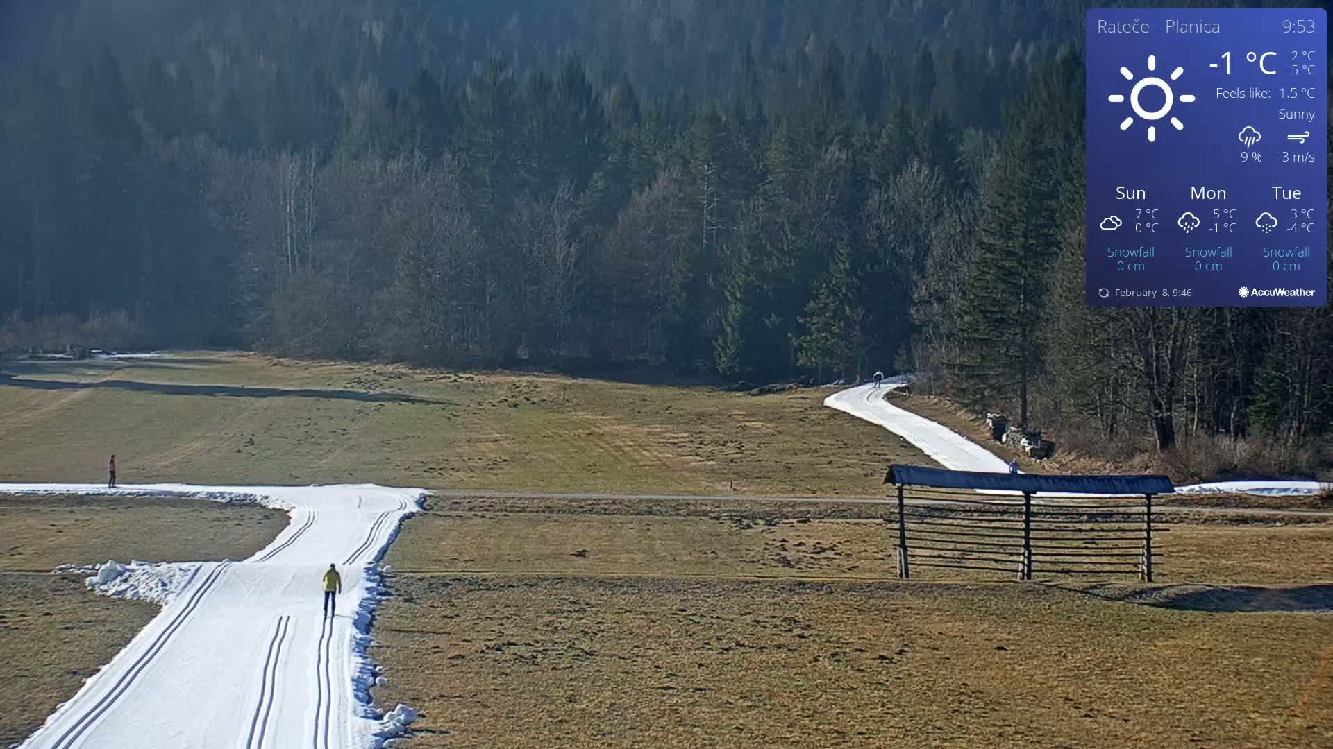 Two cross-country skiers are on a snowy track that winds through a sunny, grassy field bordered by a dark forest.
